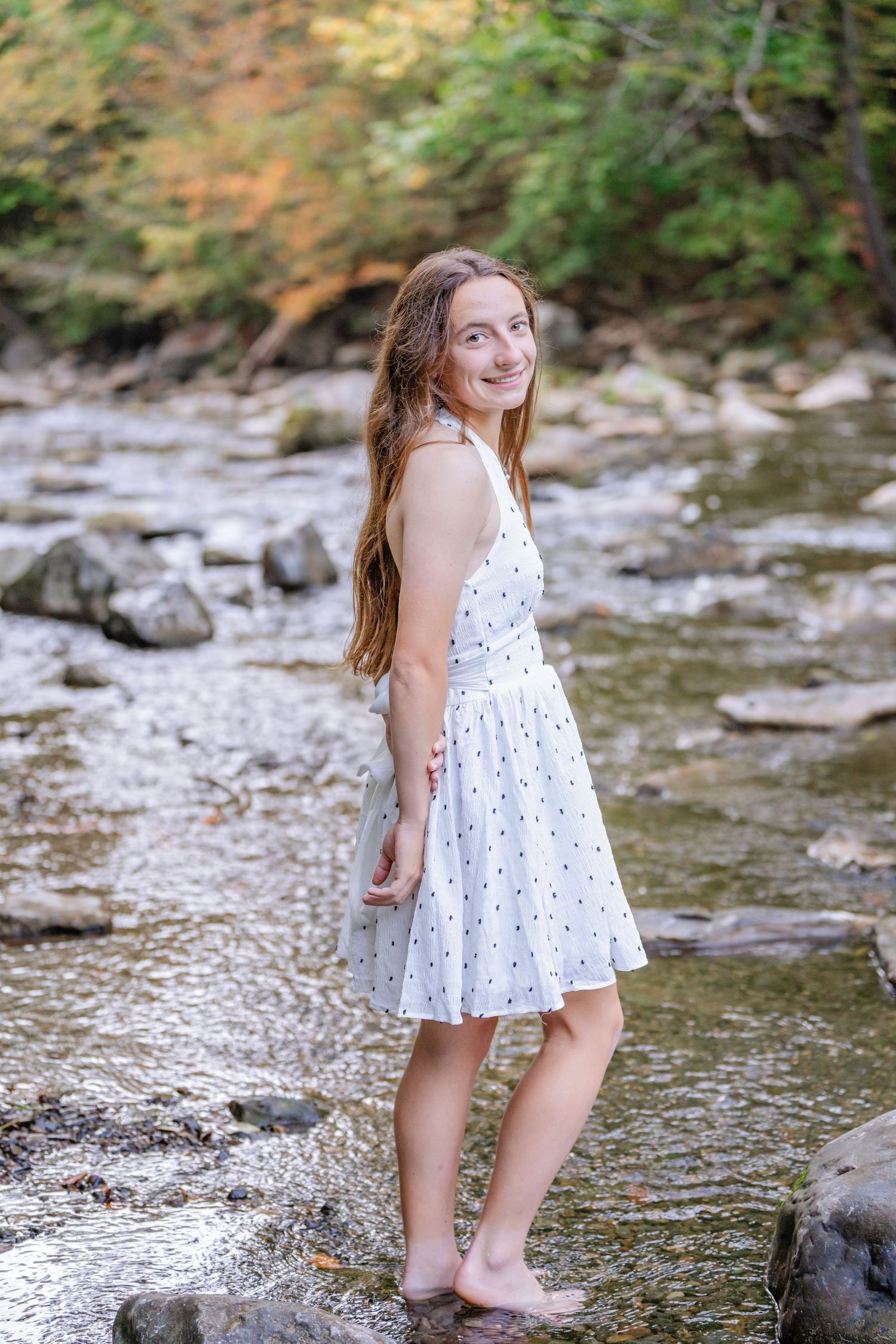 A young girl in a white polka dot dress plays by a rocky stream on a sunny summer day.