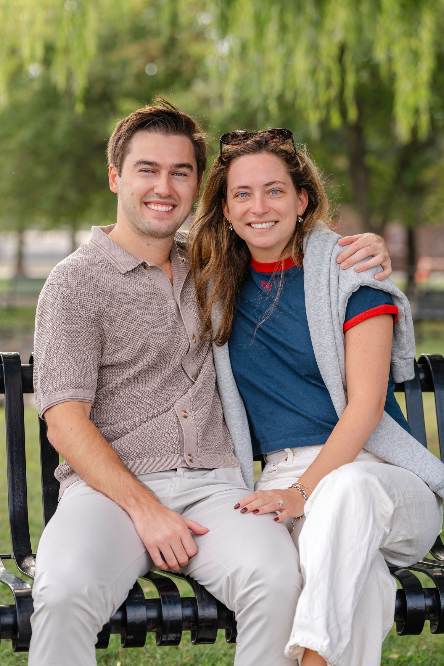 A couple sits together on a park bench surrounded by willow trees on a sunny day.