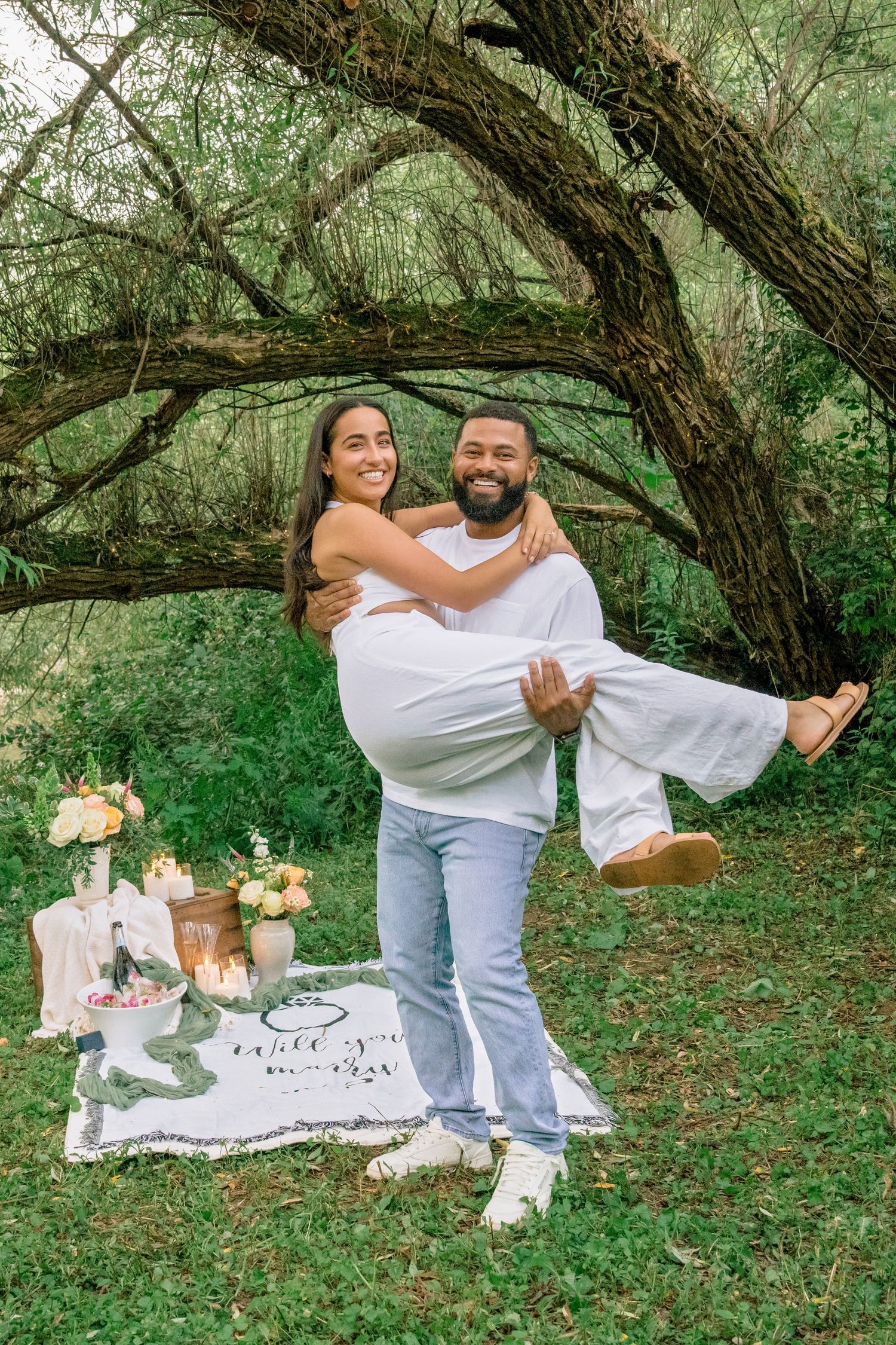 Playful couple having fun during an outdoor photoshoot near a curved tree in a wooded area.
