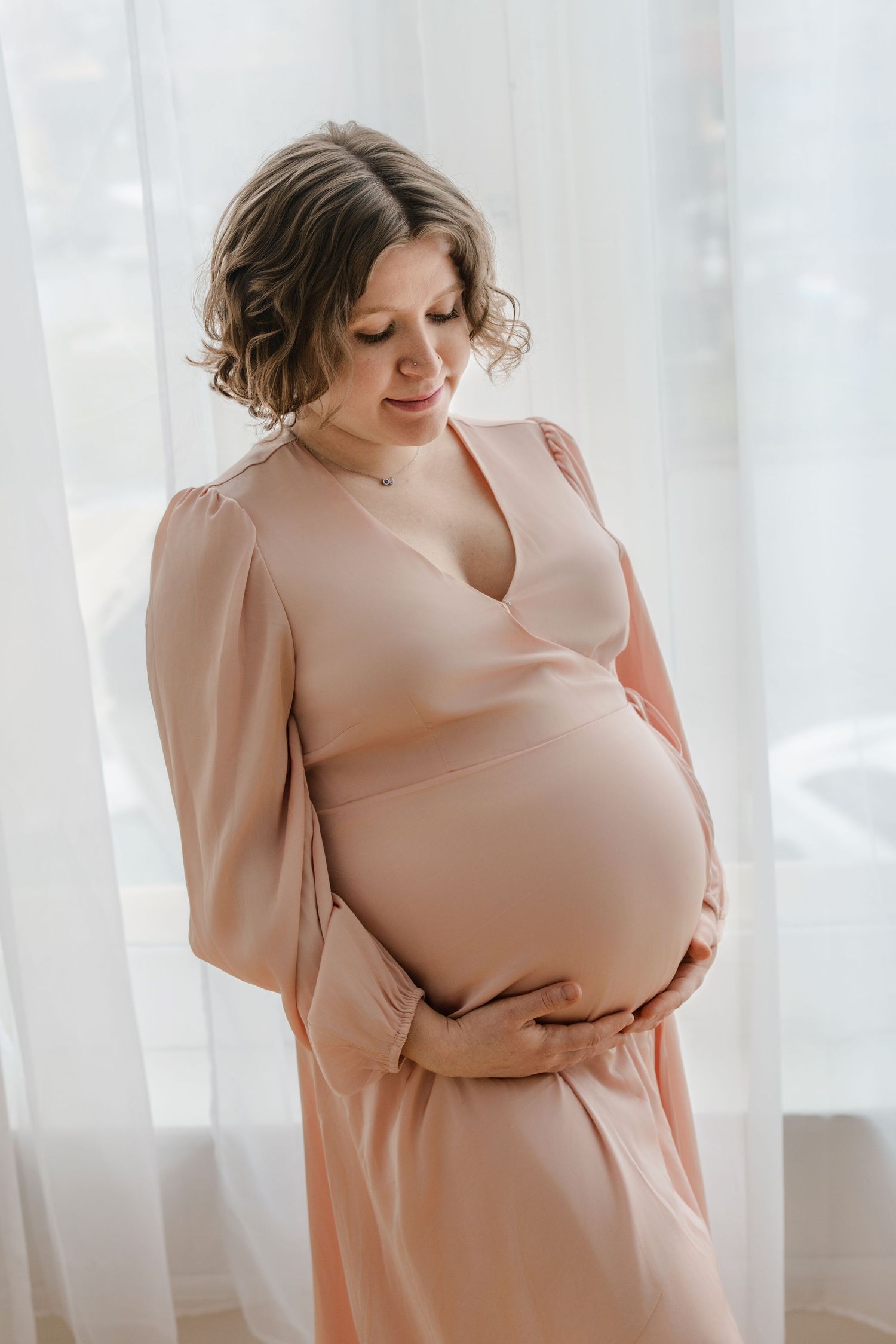 Soft, dreamy maternity portrait showing expectant mother in pink dress standing near sheer curtains.