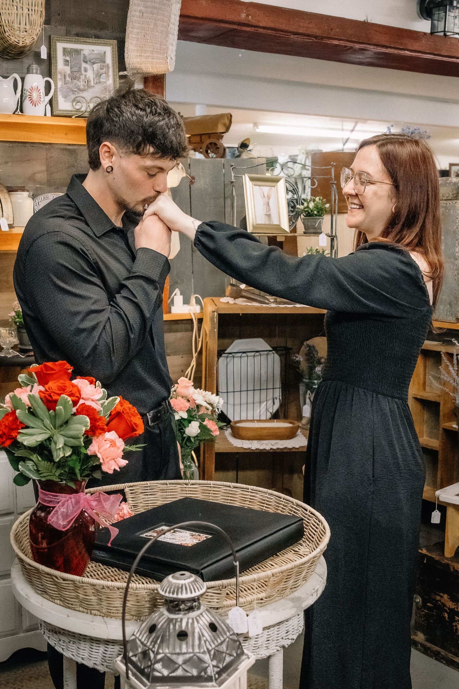 A man in black kisses a smiling woman's hand in a charming antique shop with roses nearby.