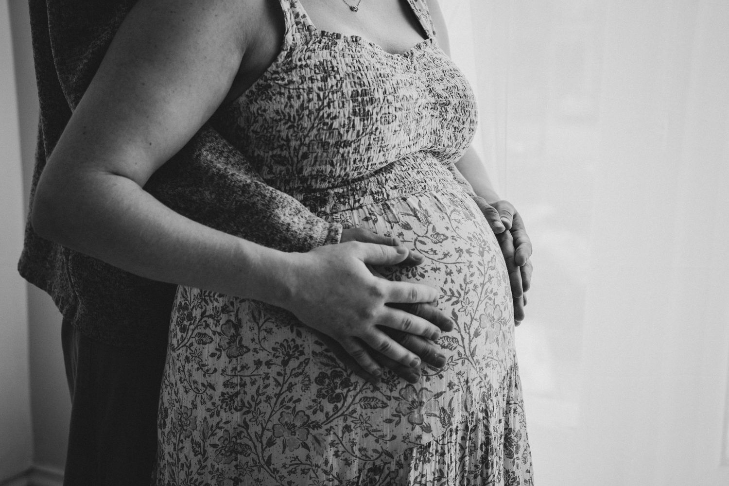 A black and white photo shows a pregnant woman in a lace dress cradling her baby bump against a light background.