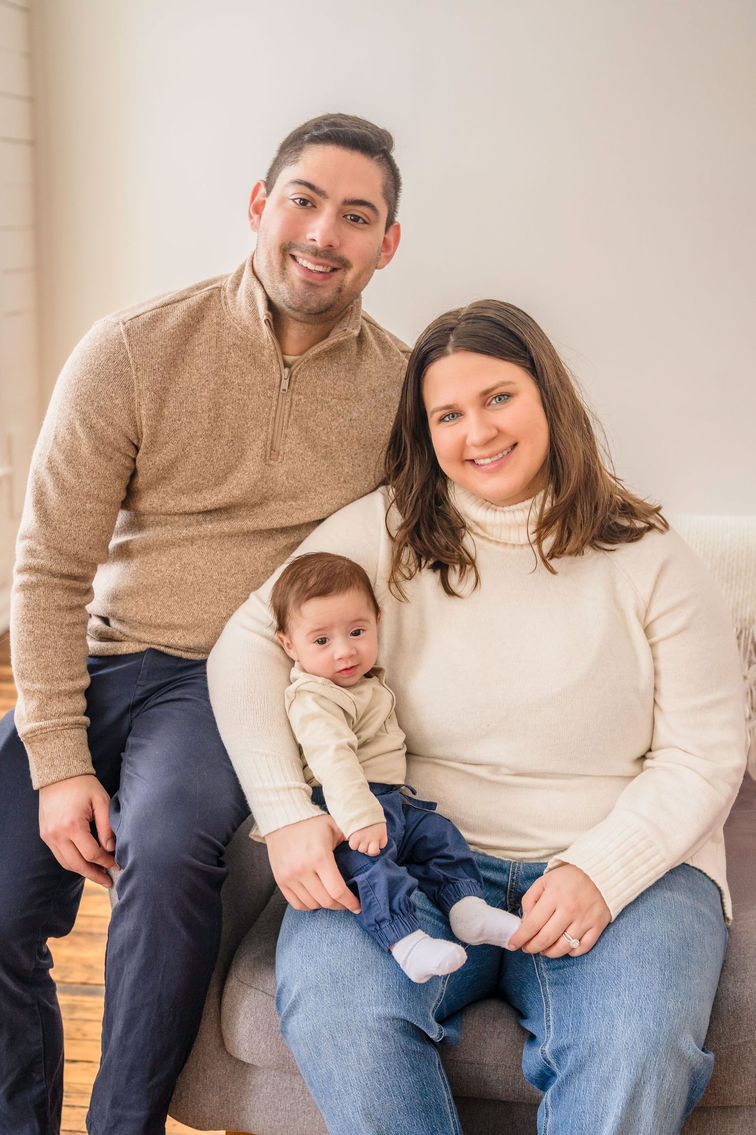 A family sits together on a couch wearing cozy cream and tan sweaters with their young baby.