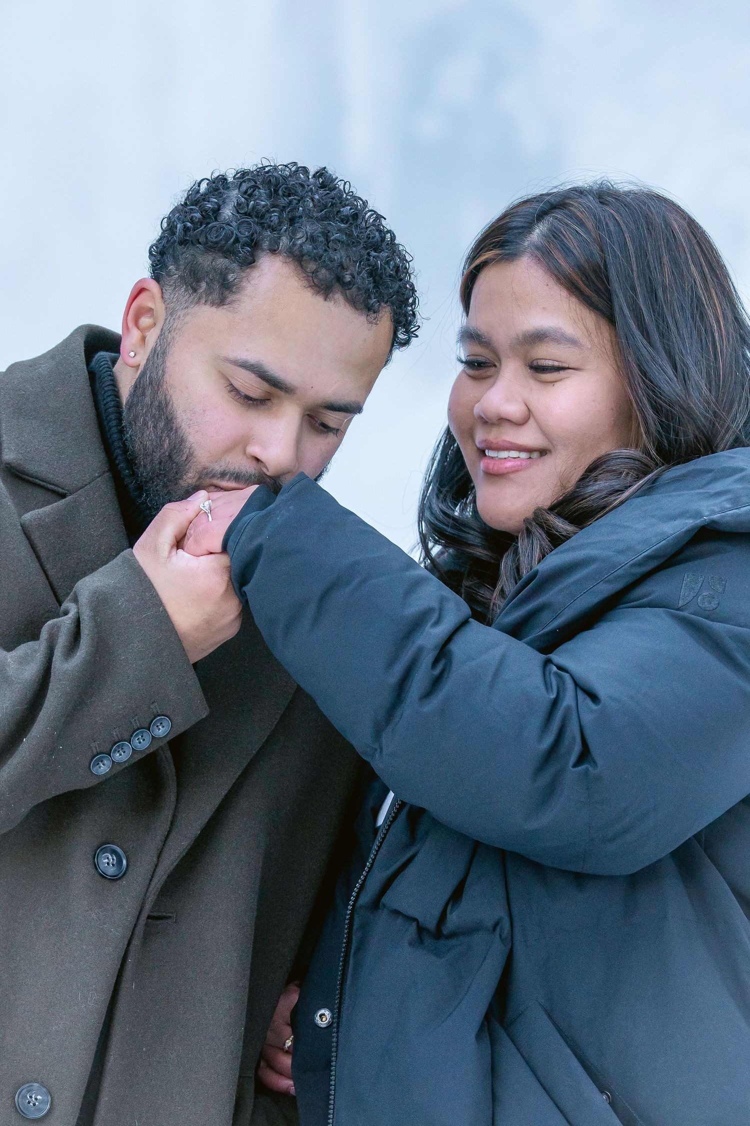 A man in a gray coat kisses a woman's hand while she smiles warmly, both dressed in winter coats outdoors.