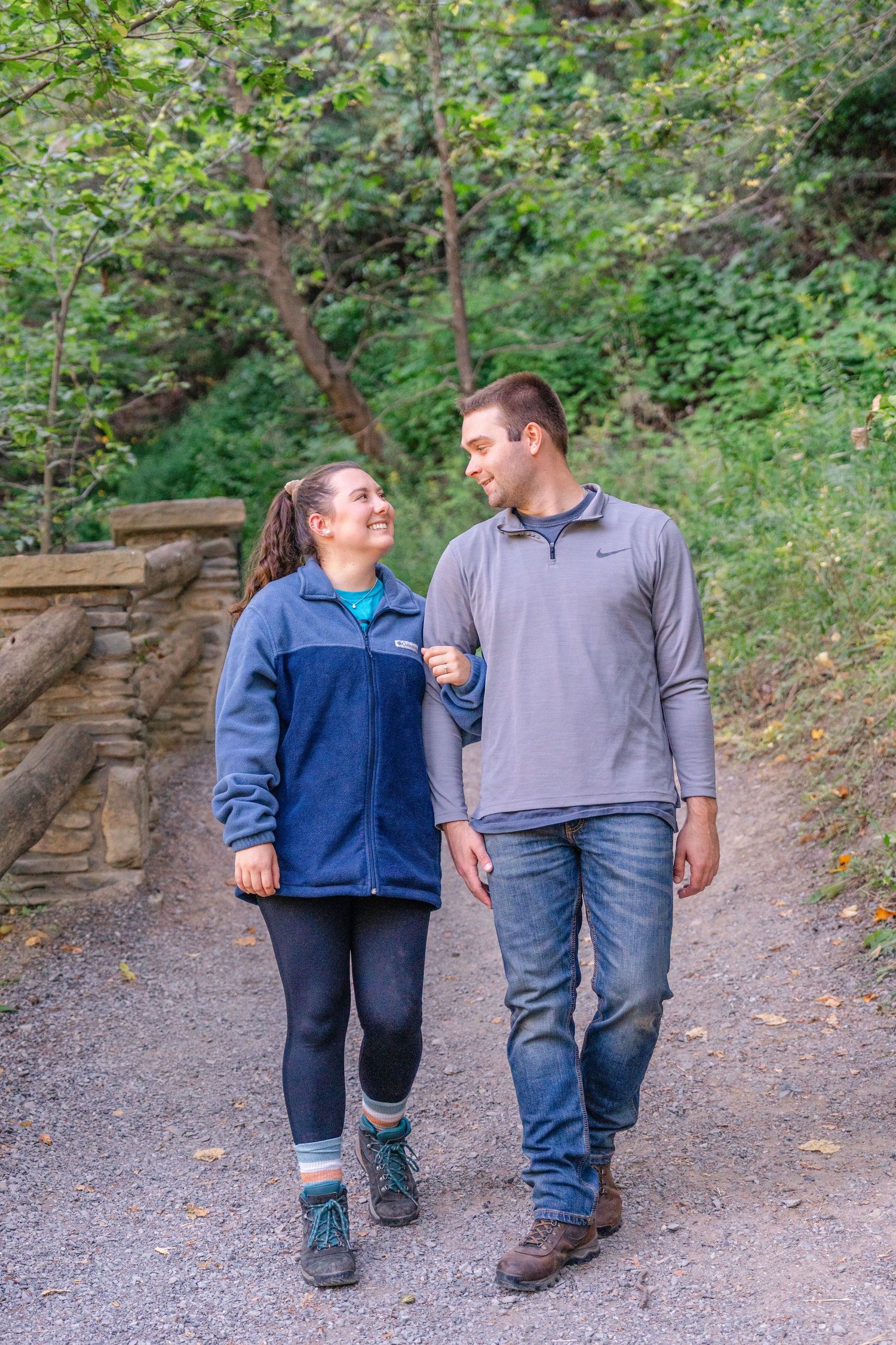 Two people take a romantic stroll together on a wooded path with stone walls visible in the background.