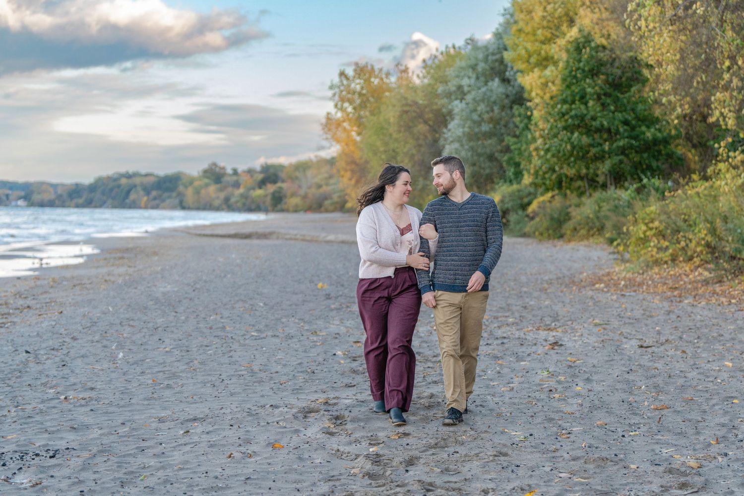 A couple walks together on a rocky beach during sunset with fall foliage in the background.