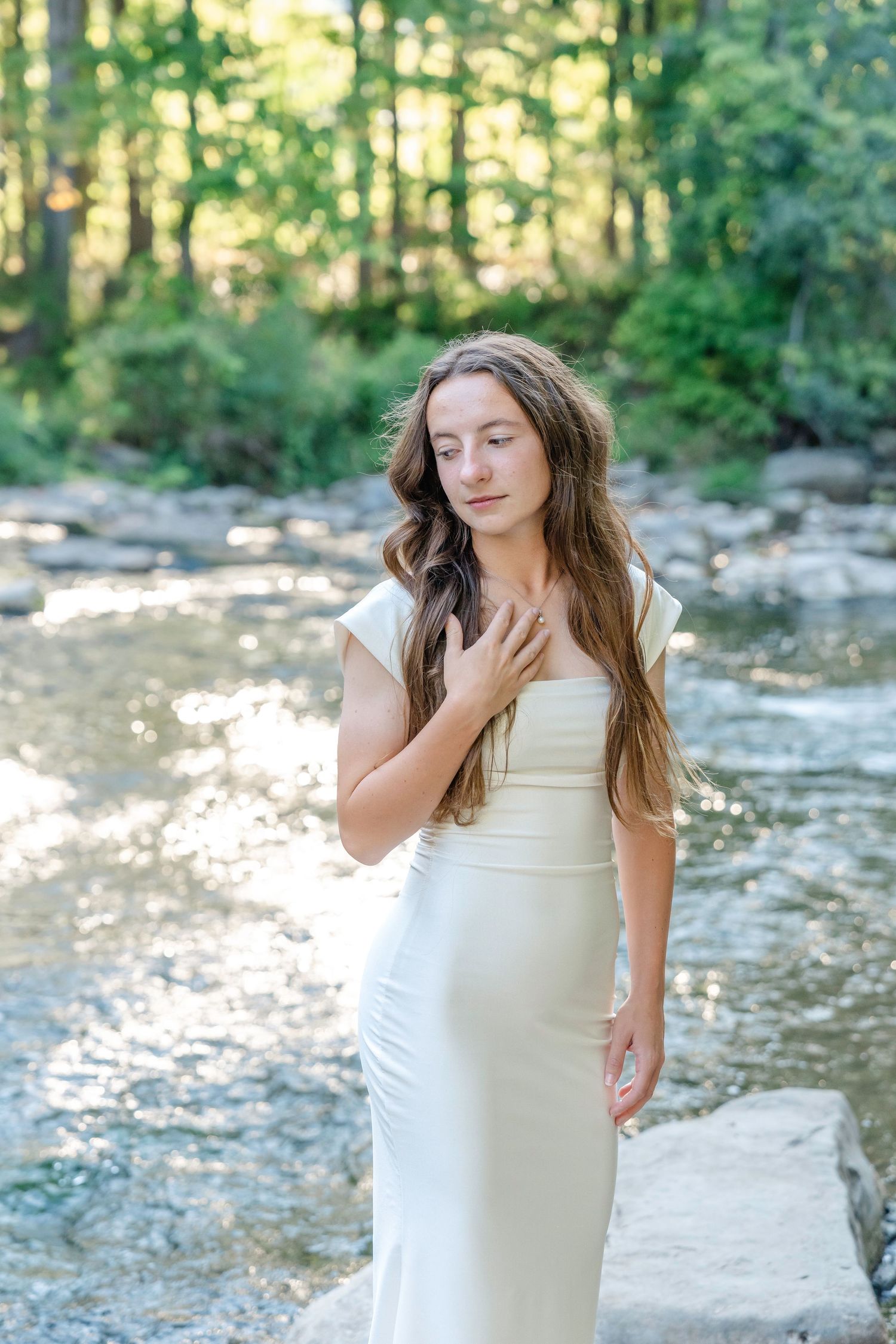 A person in a white dress stands by a rocky creek in a serene forest setting during golden hour.