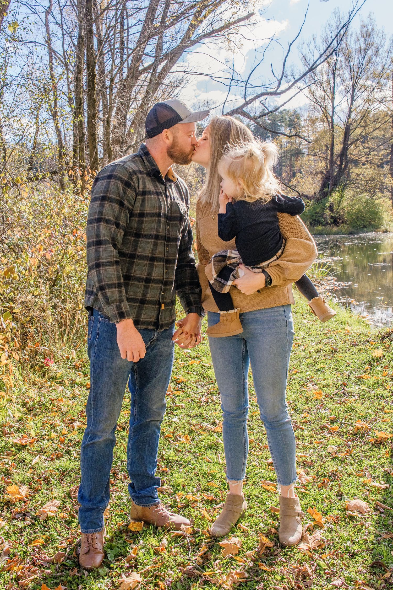 A couple in casual fall attire stands together on autumn leaves by a pond during a photoshoot.