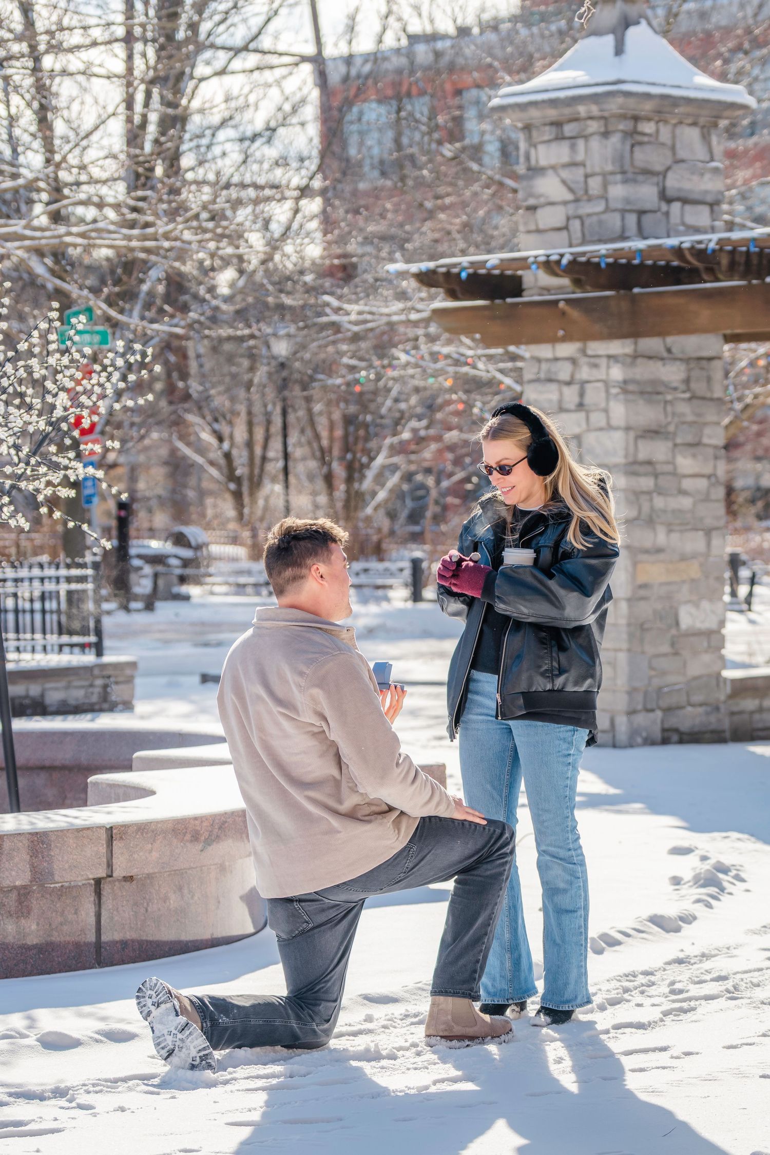 Man proposes on one knee to woman in snowy winter park, holding ring box outdoors.