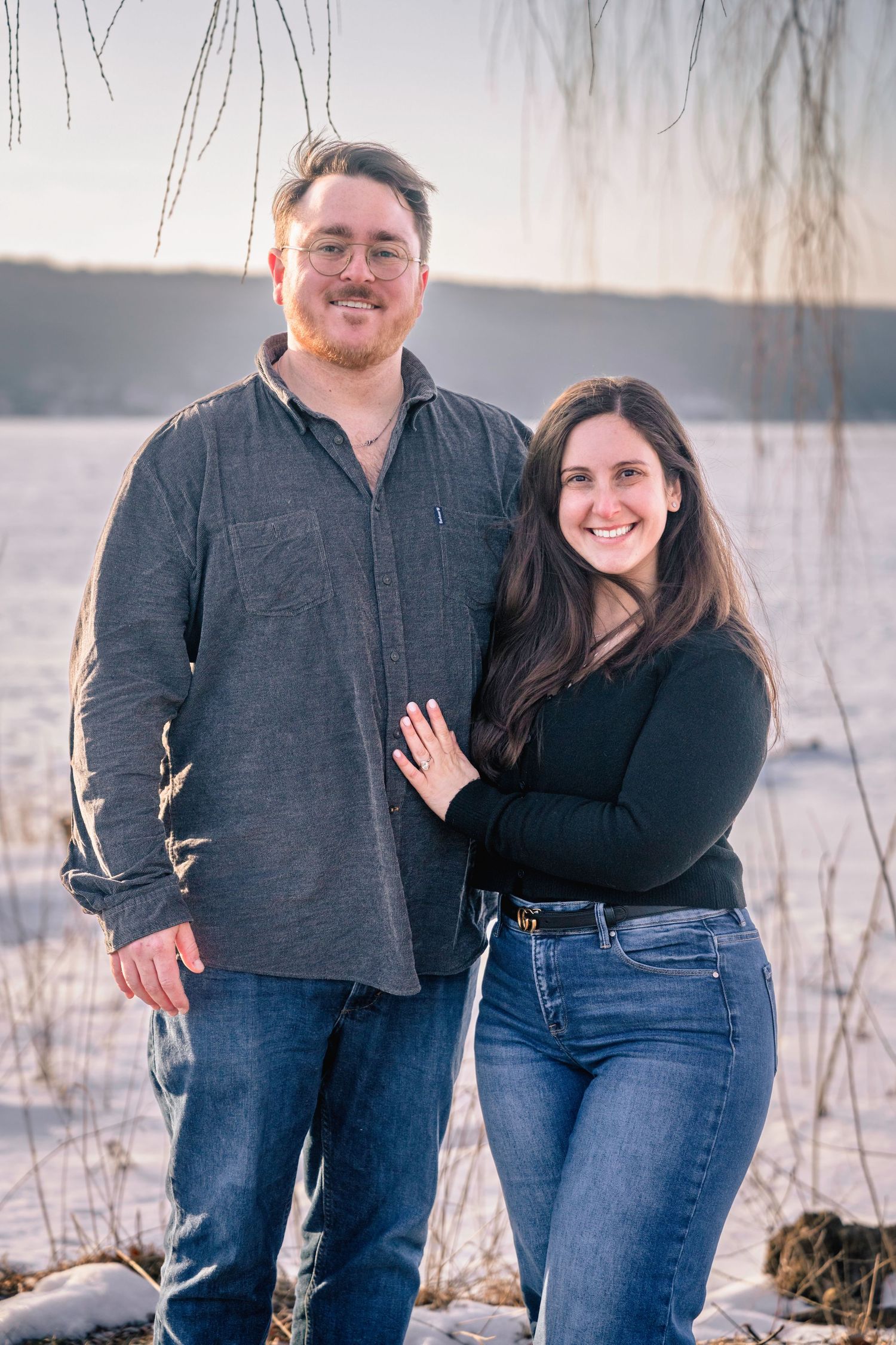 A smiling couple poses outdoors in winter, standing near a frozen lake with snow-covered ground and bare willow branches.