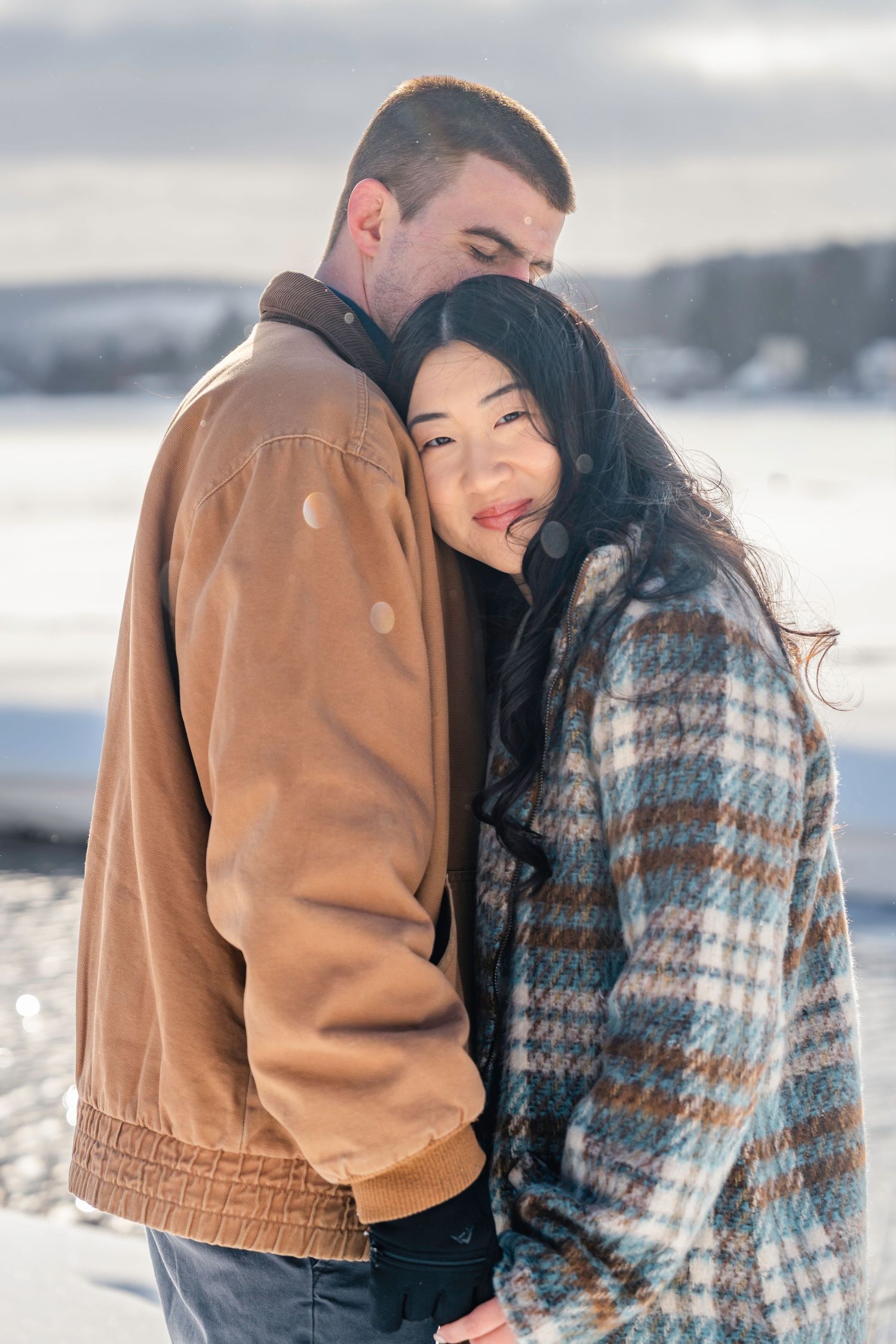 A couple embraces in a snowy winter landscape, woman in plaid coat leans on mans shoulder, golden sunlight glowing.