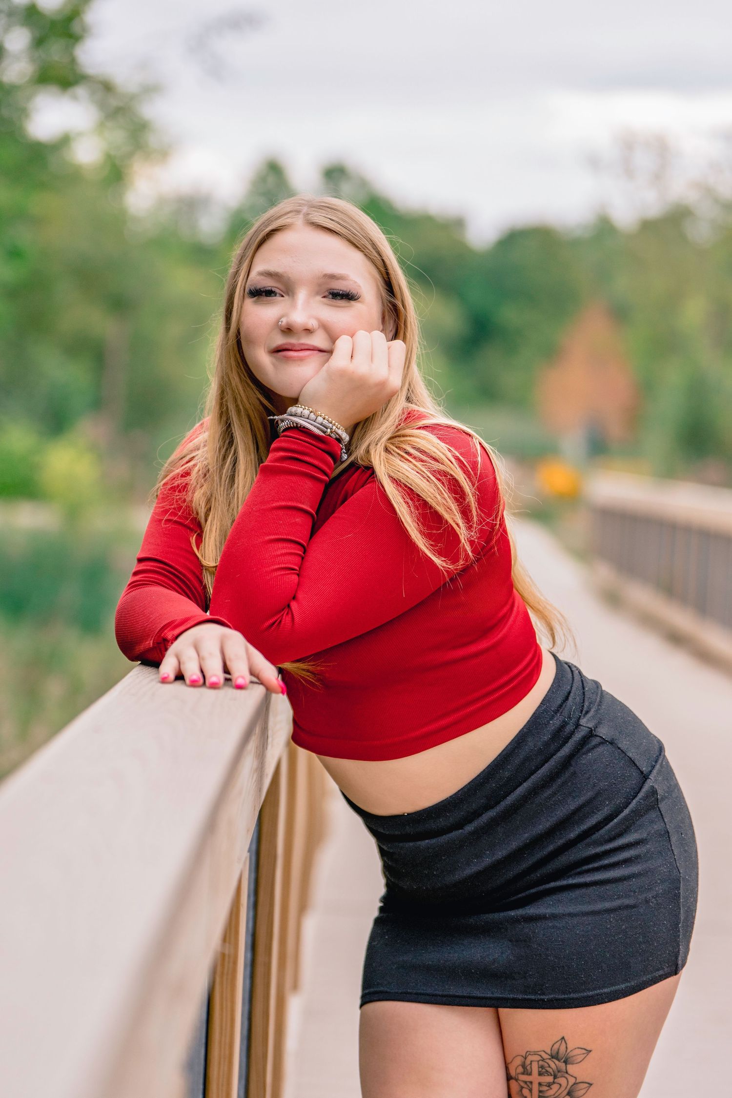 A person in a red long sleeve top and black skirt poses casually against a railing on a path outdoors. Senior portraits.