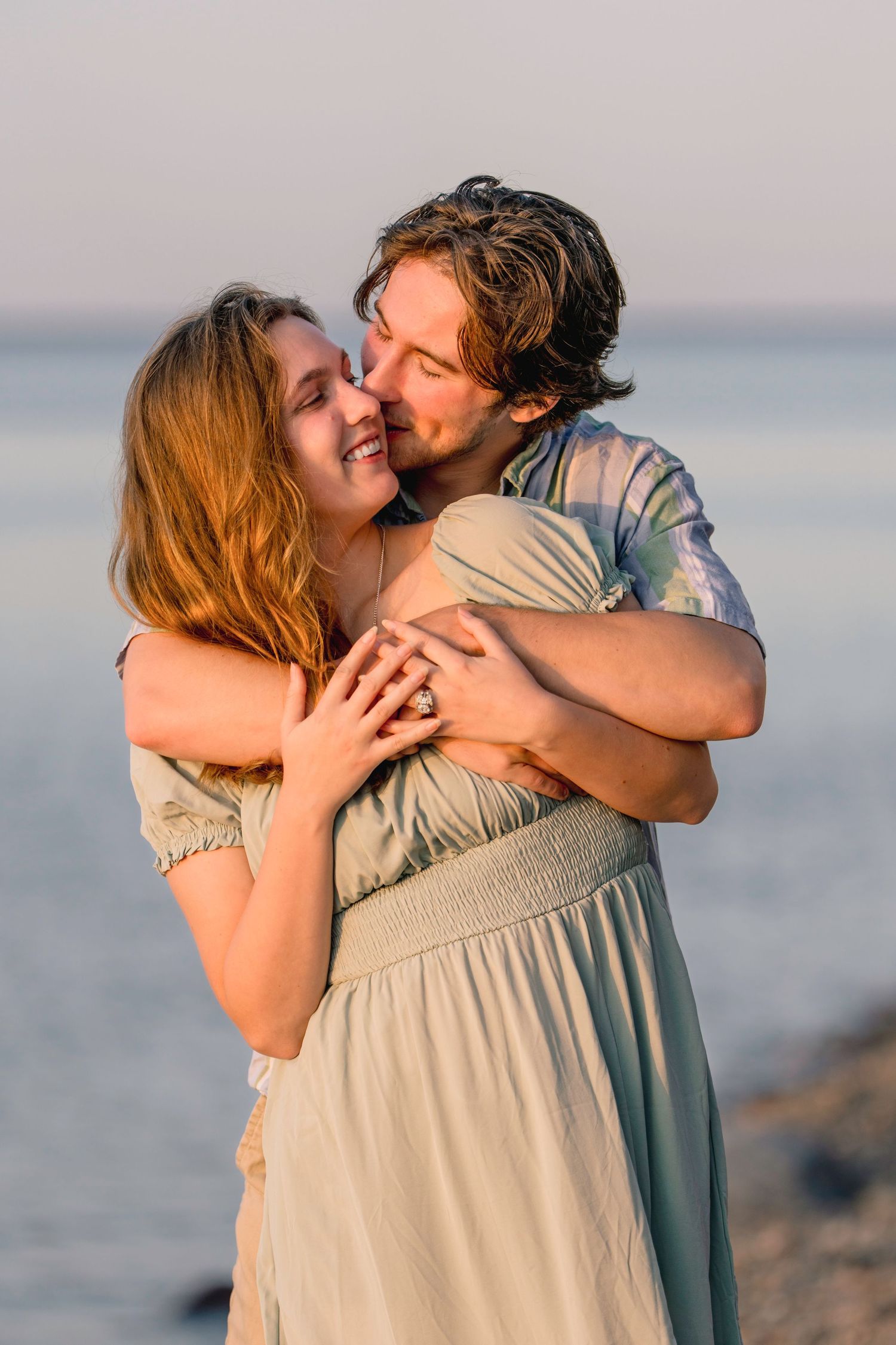 A romantic couple shares tender kisses and embraces on a beach at sunset. Chimney Bluffs State Park proposal and engagement session.