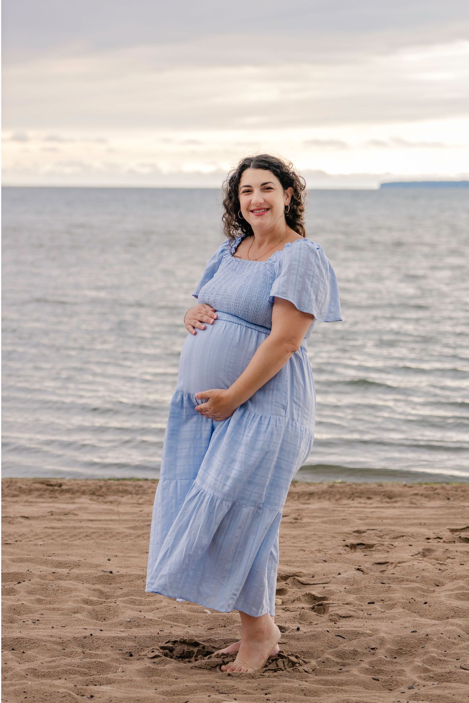 A person in a light blue dress poses on a sandy beach at sunset, cradling their baby bump.