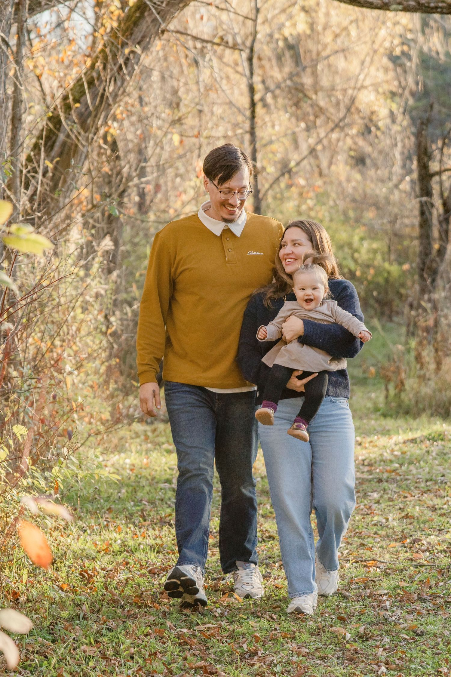 A family walks together through fallen autumn leaves in a sunlit forest setting.