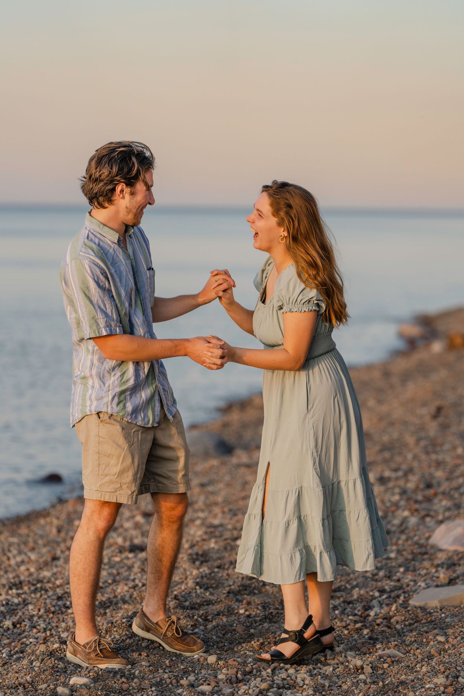 Two children dance and play together on a pebble beach with the ocean and sunset in the background.
