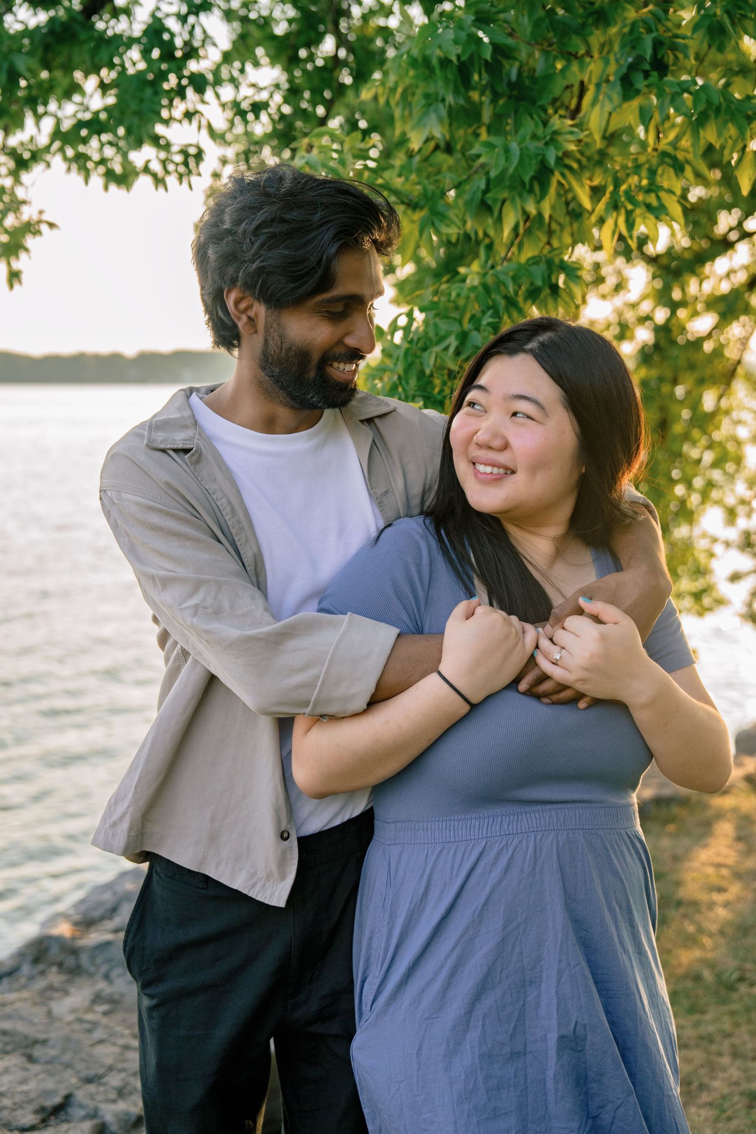 A couple embraces and laughs together outdoors with green foliage in the background during a sunny day.