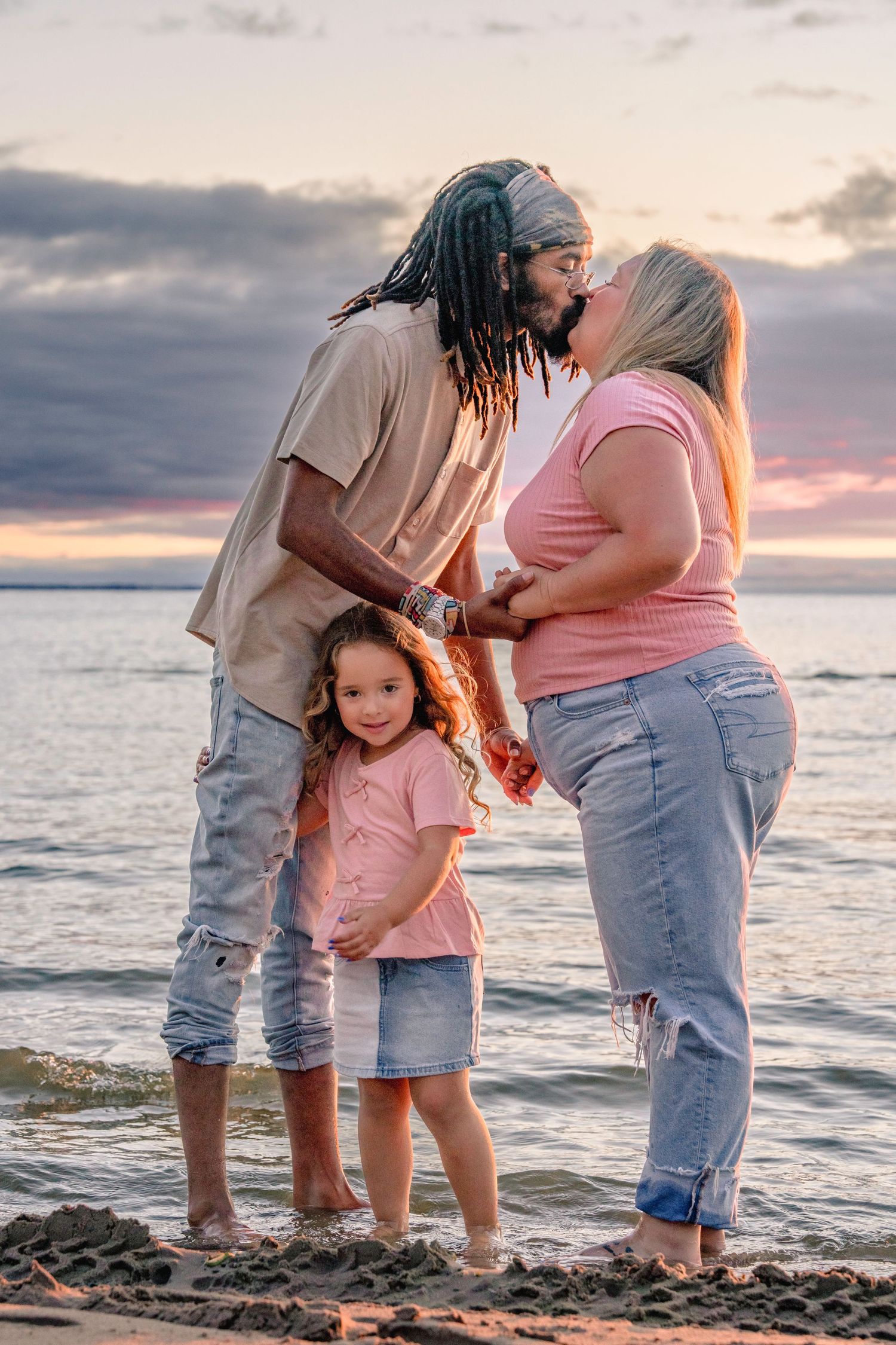 Family shares tender moments during sunset photoshoot at beach with ocean and cloudy sky in background.