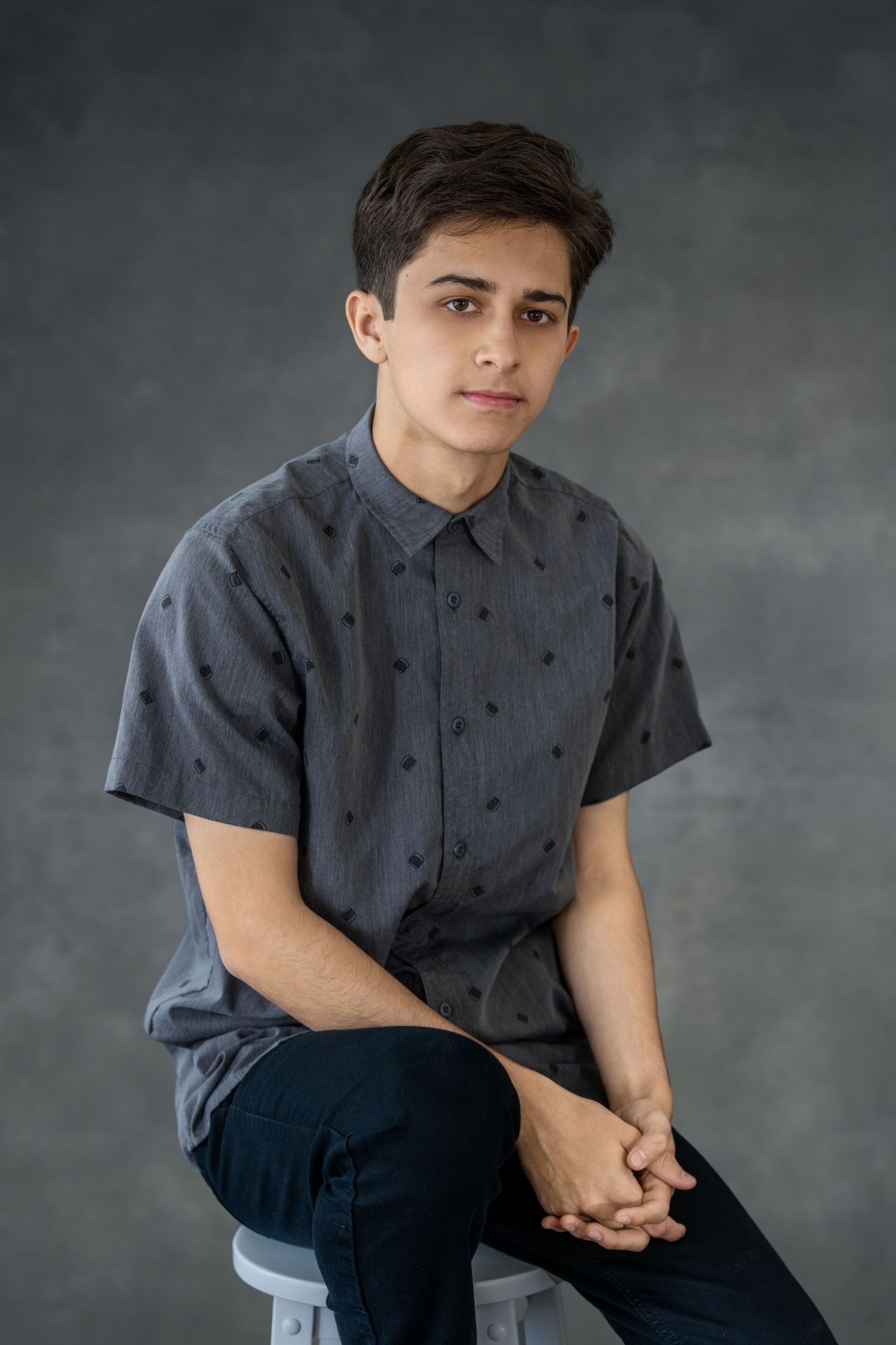 Young person wearing a dark button-up shirt poses against a gray background in a studio setting.