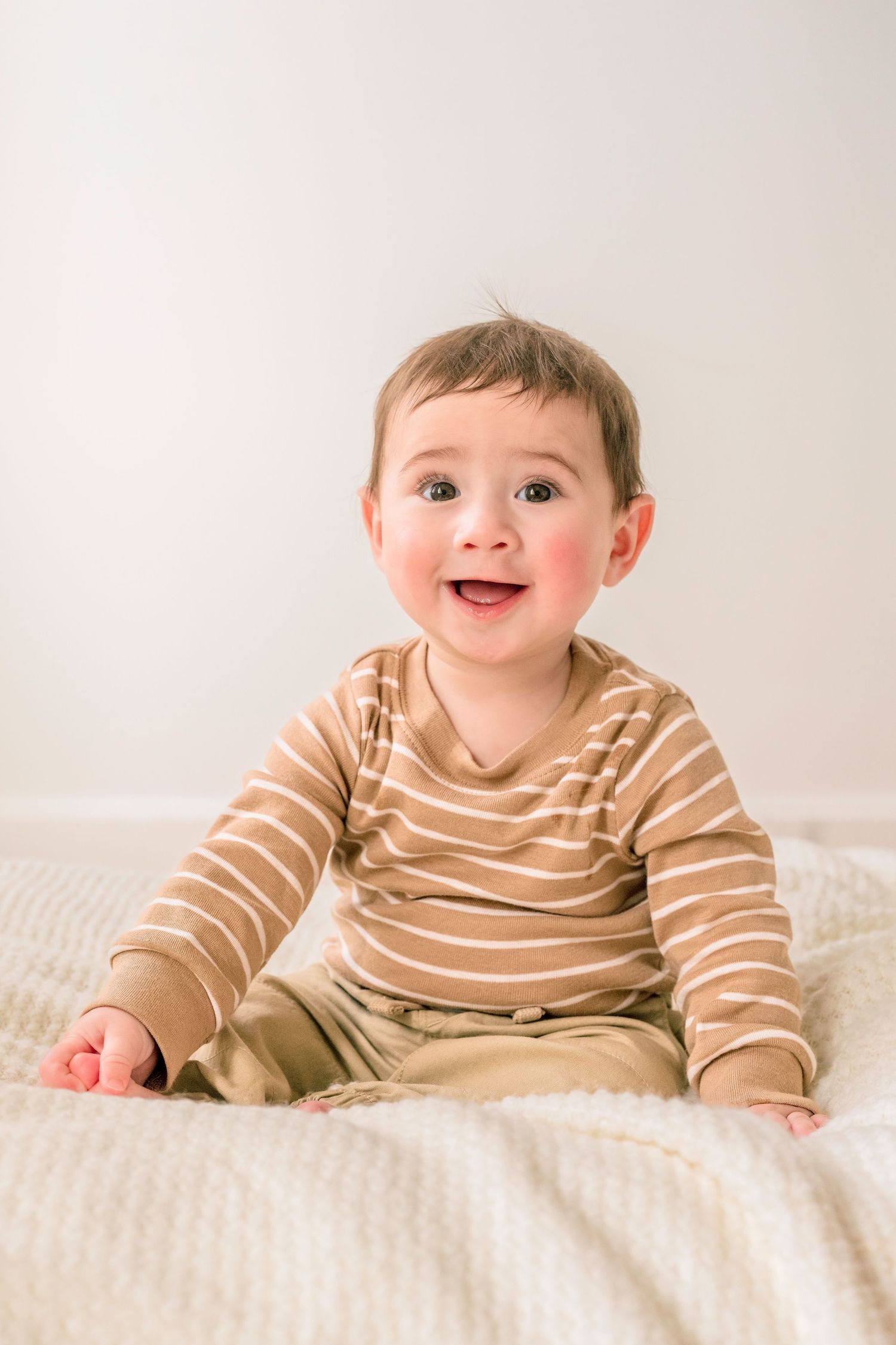 Smiling baby in striped brown outfit sitting on white blanket.