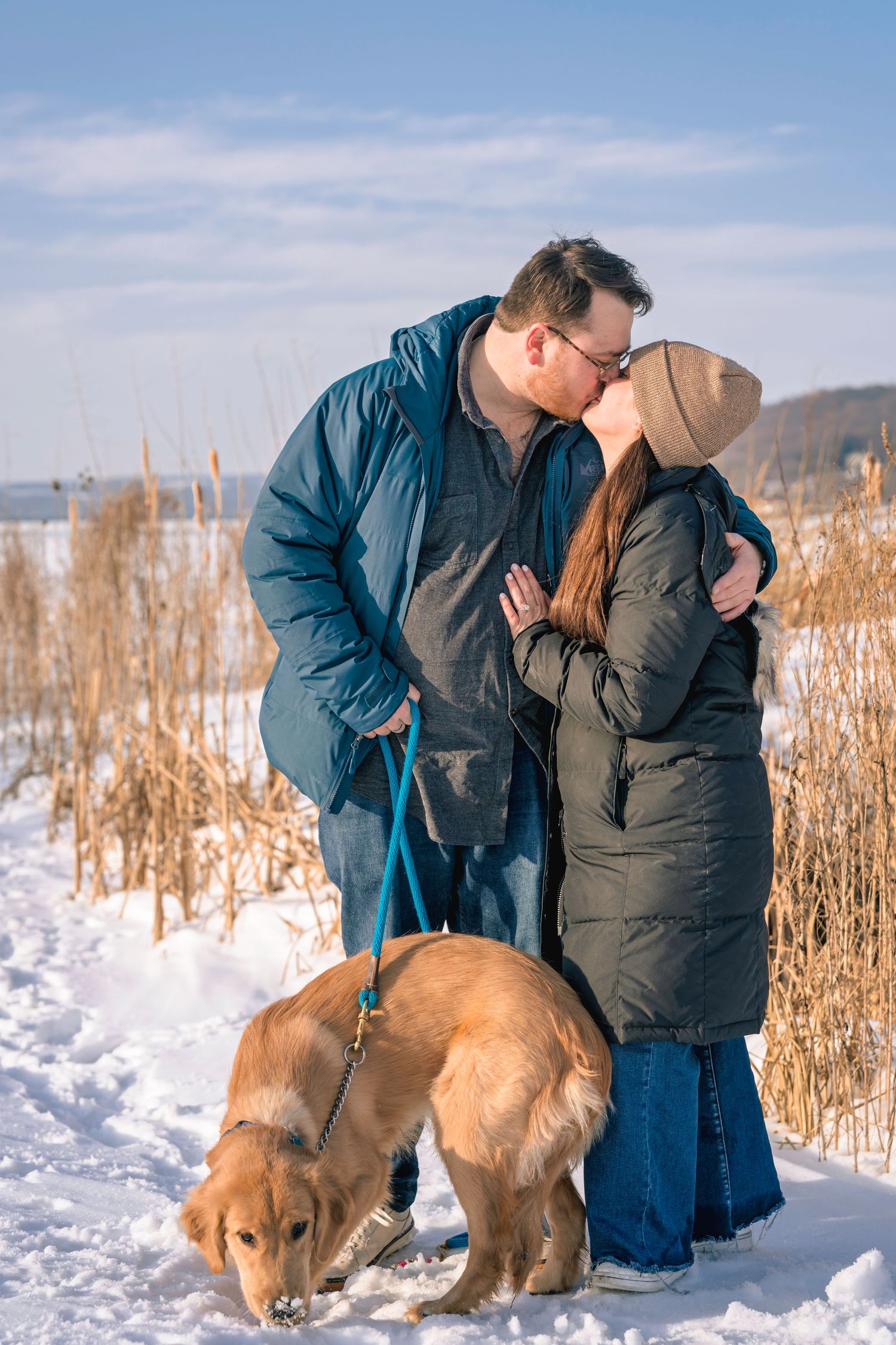 Couple kissing outdoors in winter snow with golden retriever on leash near dry reeds on sunny day.