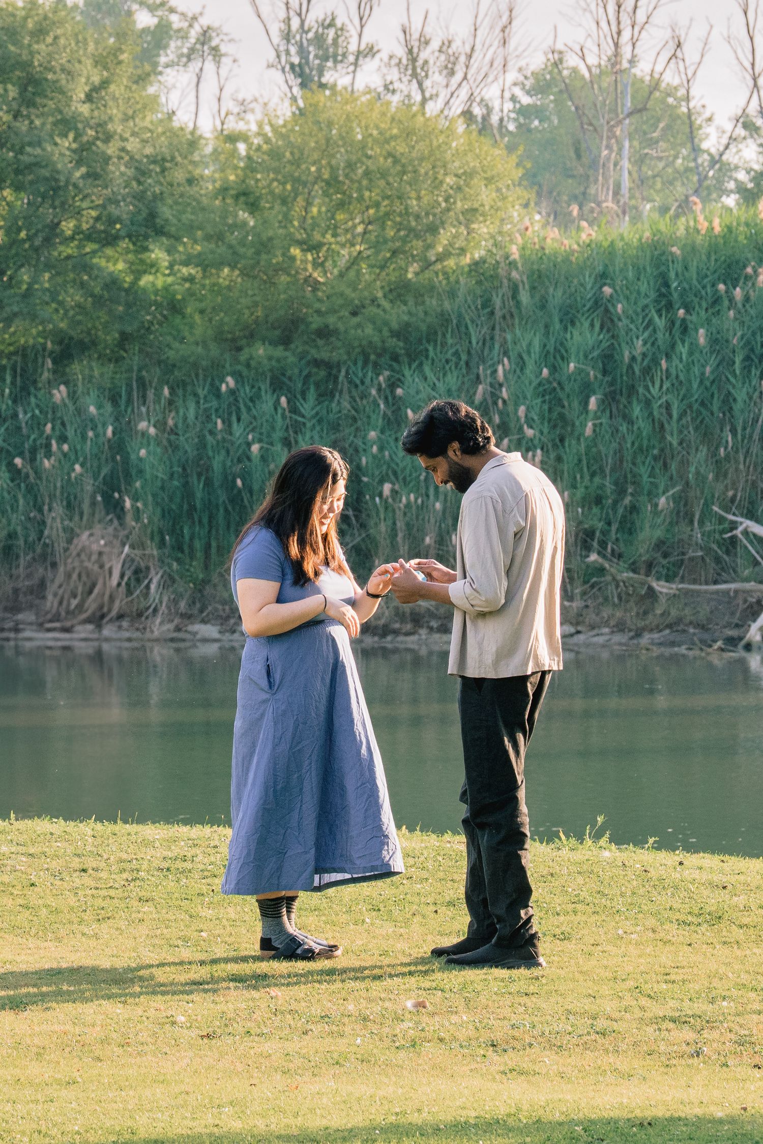 A couple stands on grass by a lake during a romantic proposal moment at sunset.