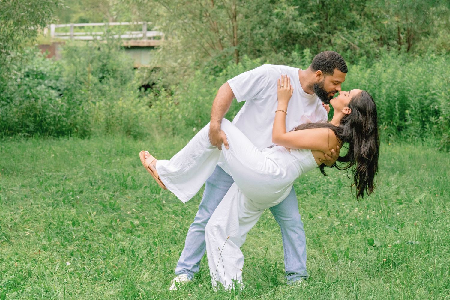 A couple in white outfits shares romantic moments in a green meadow during a summer photo session.