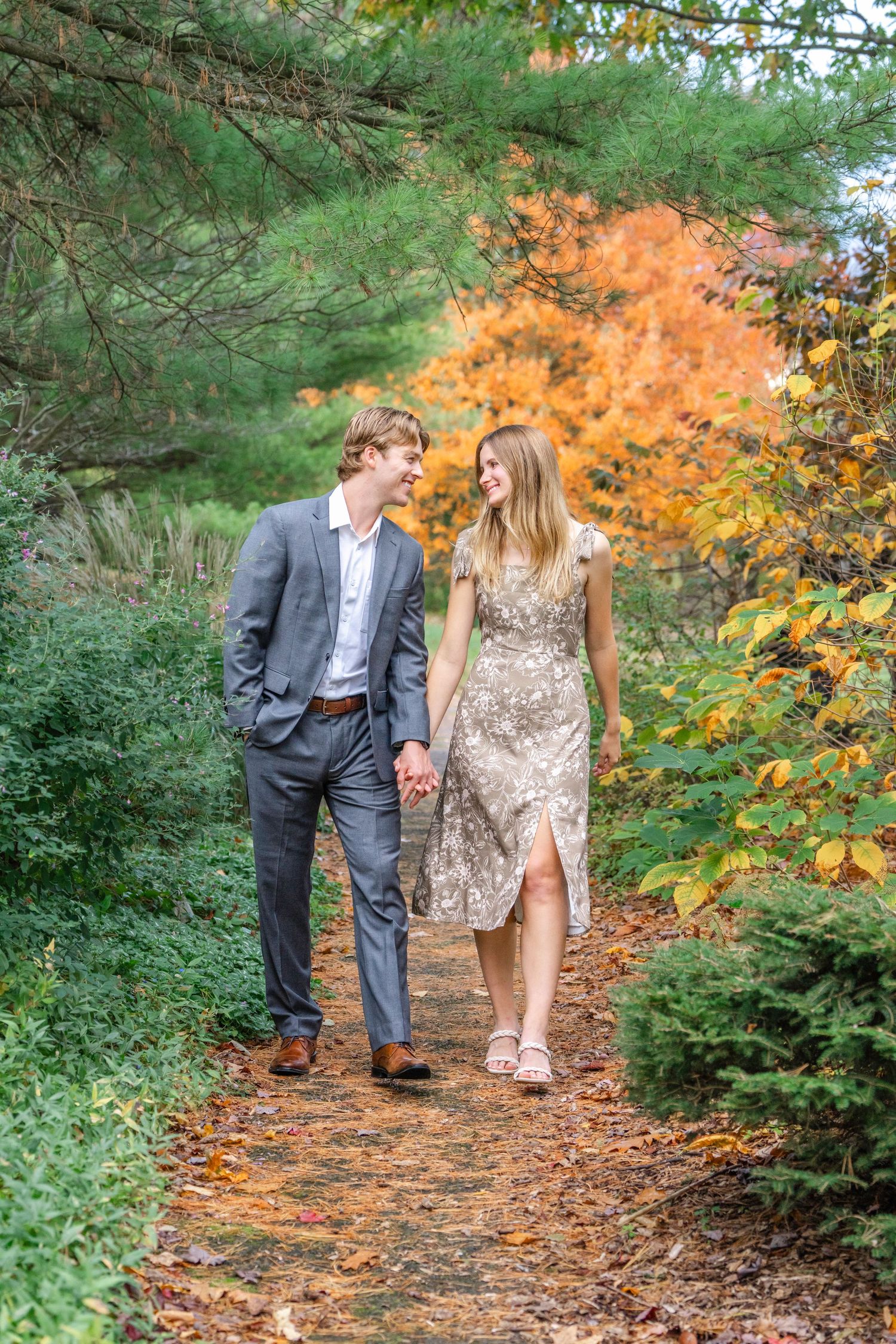 A couple walks hand in hand along a scenic autumn forest path surrounded by colorful fall foliage and evergreen trees.