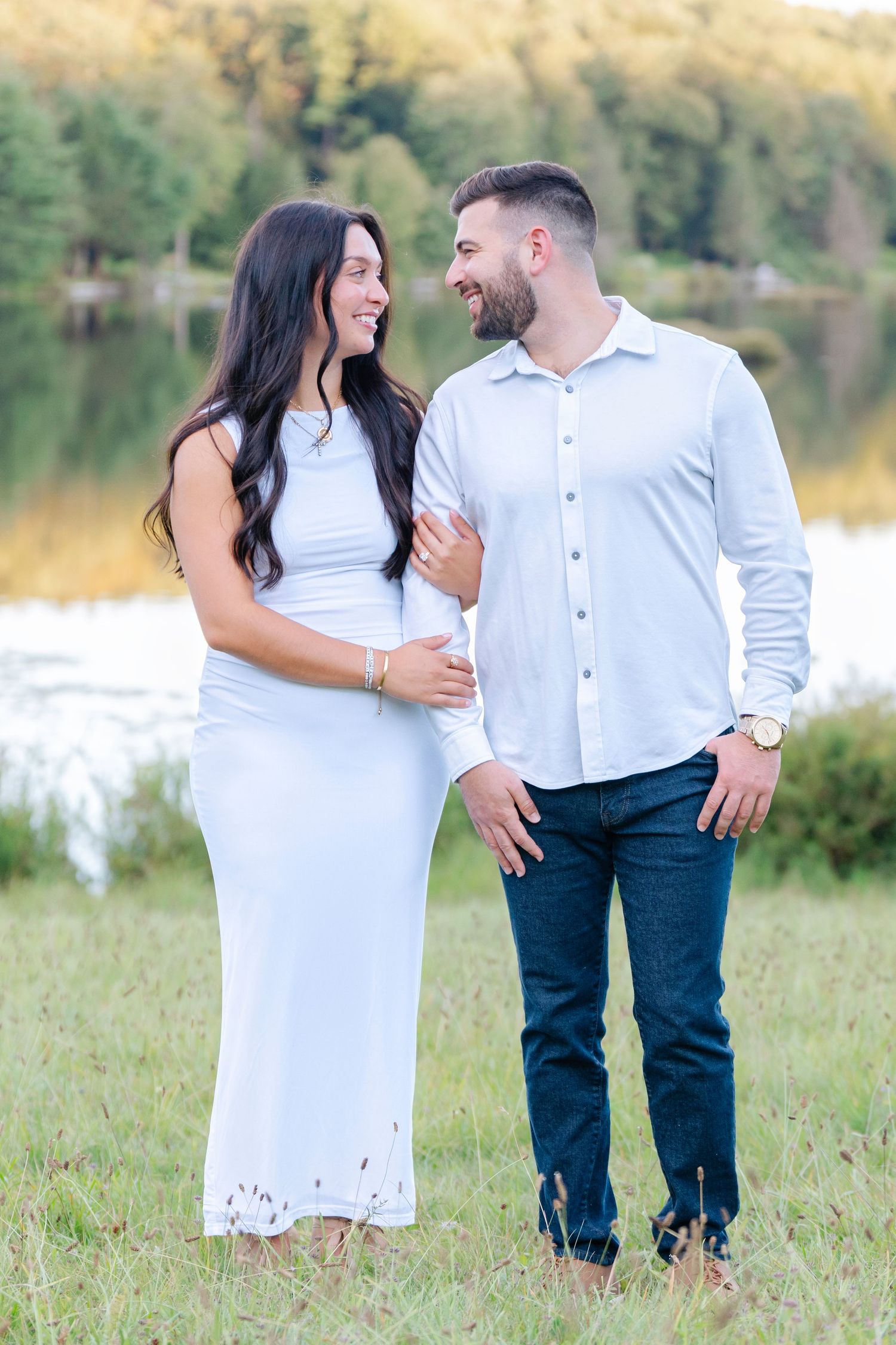 Happy couple laughs together during sunset photoshoot by a lake wearing matching white tops.