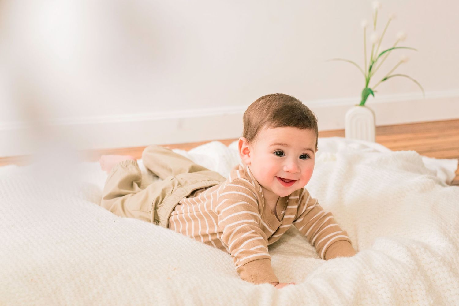 Smiling baby in striped onesie lying on white bedding with a bright airy background.