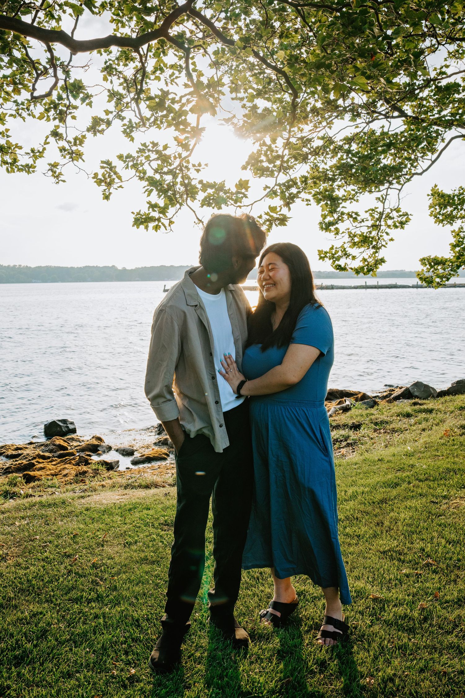 Couple sharing a romantic moment by the lake at sunset under tree branches with sunlight streaming through the leaves.