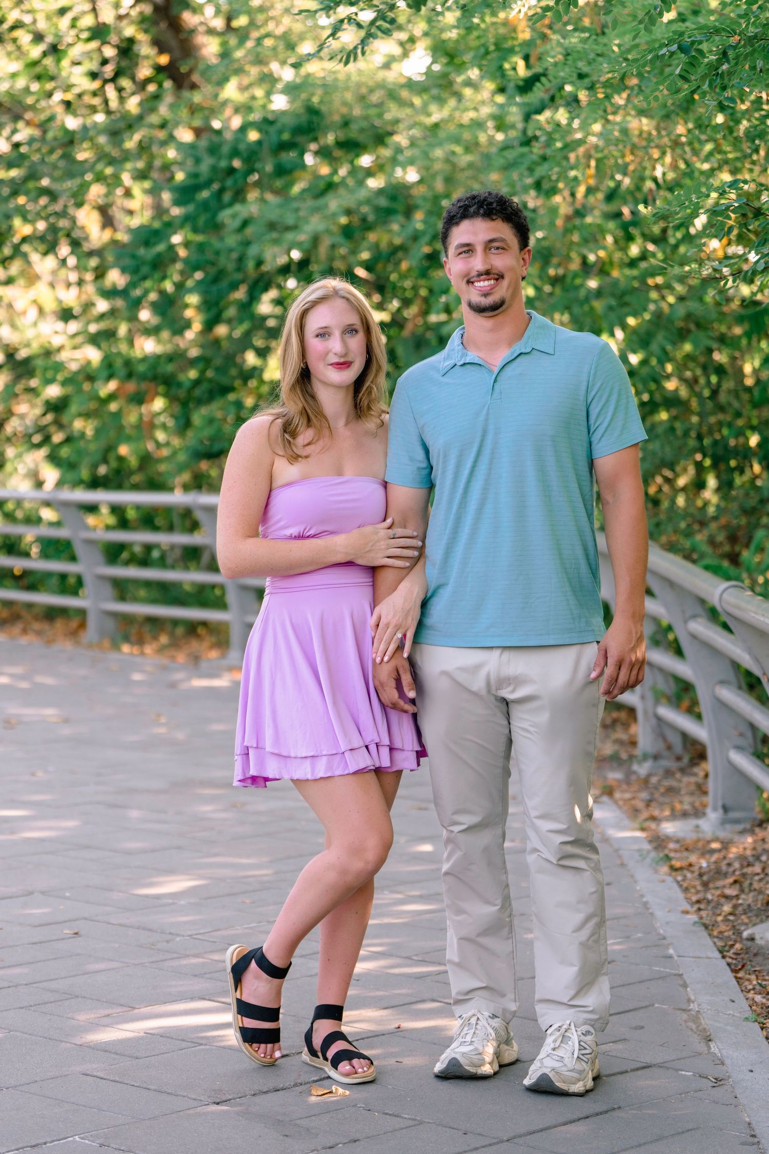 A couple in coordinated outfits poses playfully on a walkway surrounded by greenery.