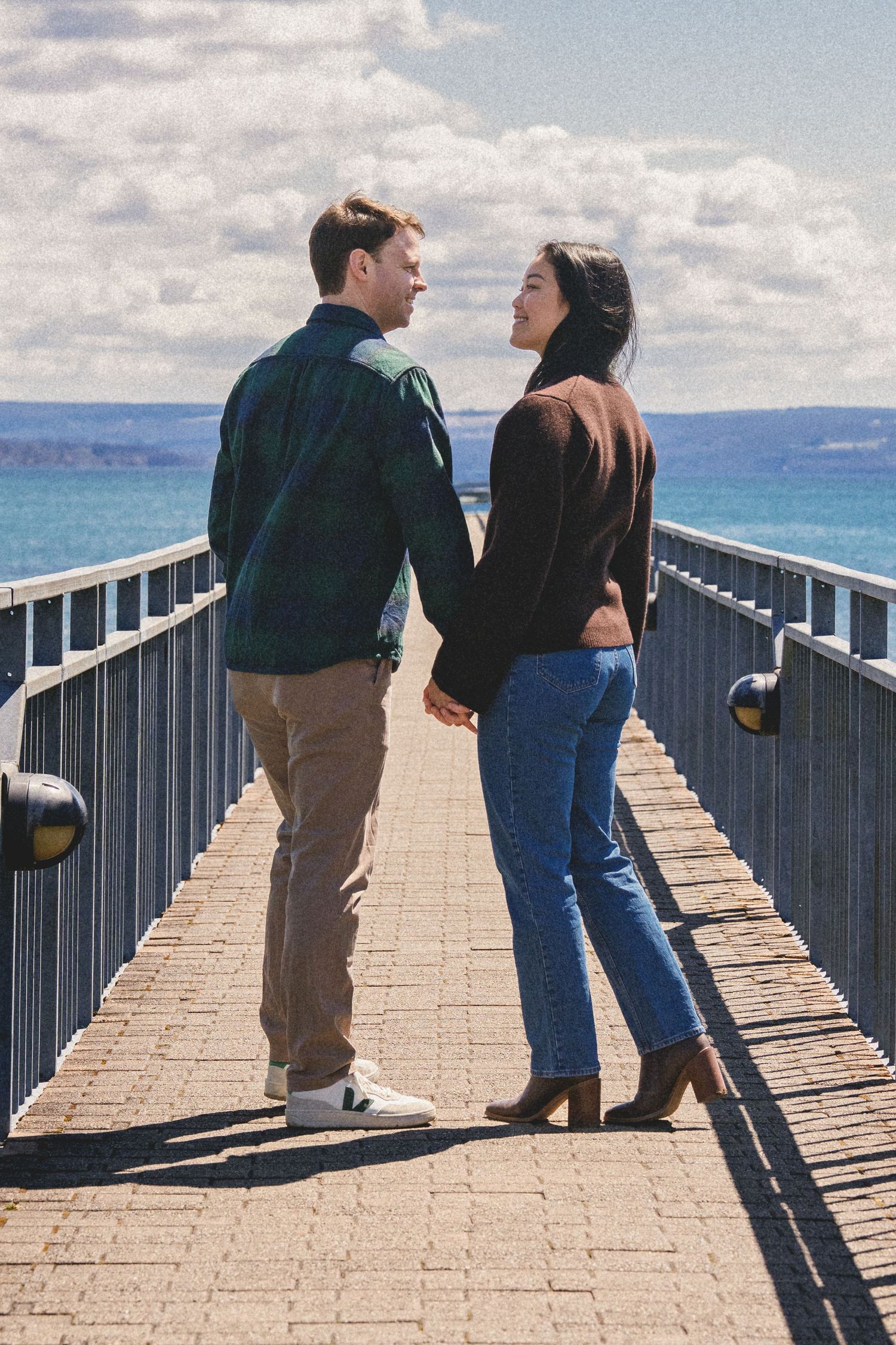 A couple walks and poses together on a wooden boardwalk overlooking the ocean on a sunny afternoon.