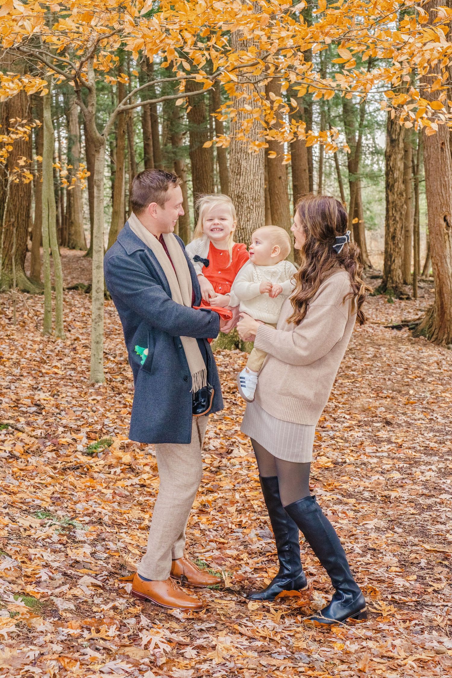 A family stands together on fallen autumn leaves in a forest with golden foliage overhead.