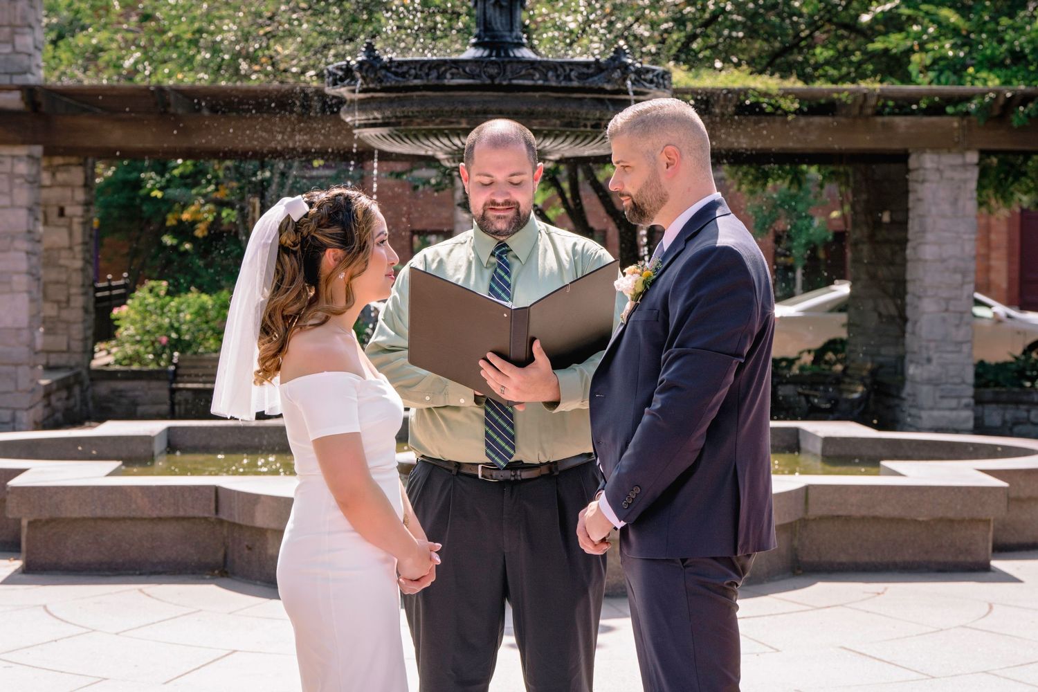 A wedding ceremony takes place outdoors on a sunny day with stone architecture and greenery in the background.