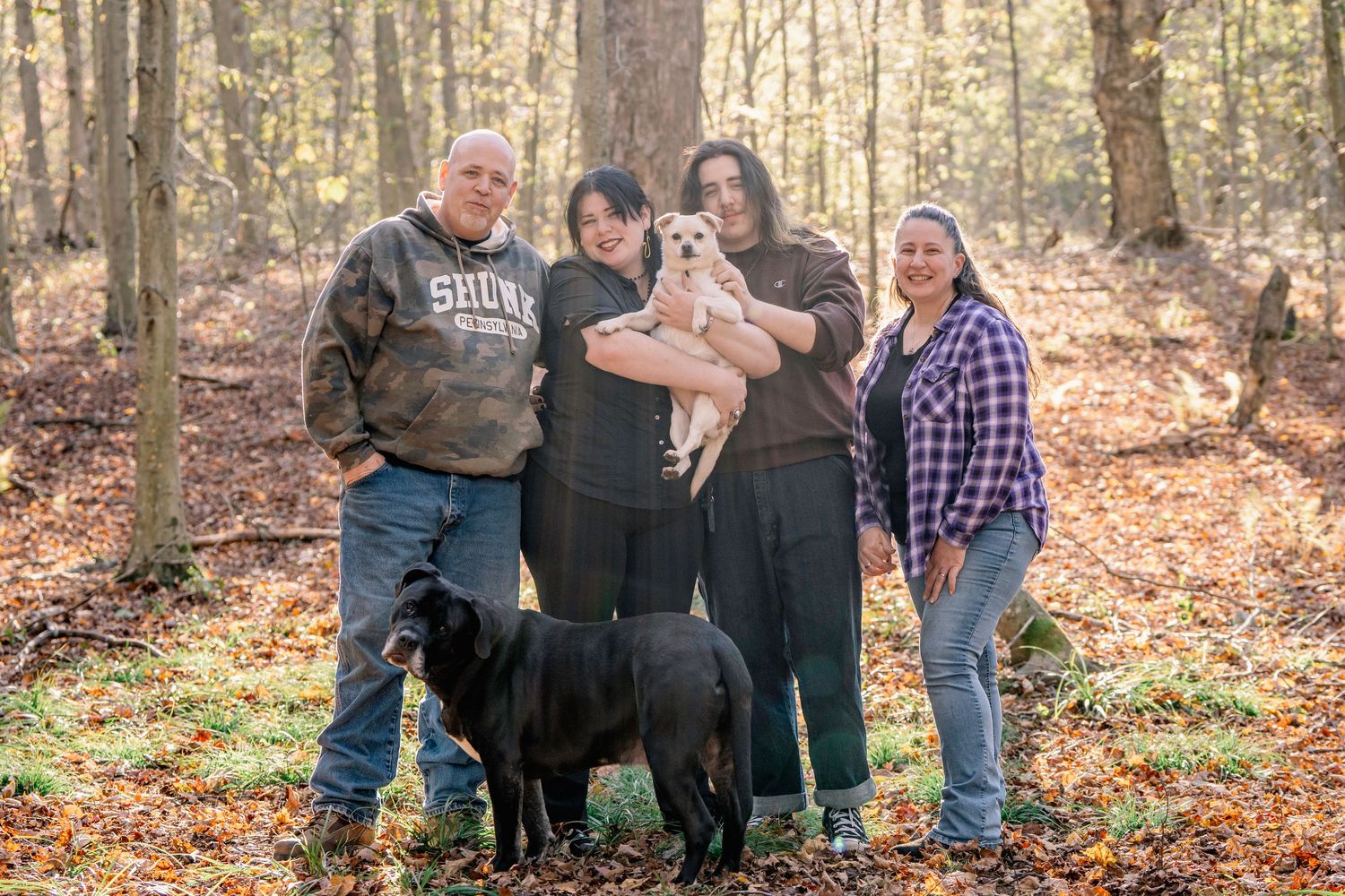 A group stands together in an autumn forest with a black dog on fallen leaves during a family photoshoot.