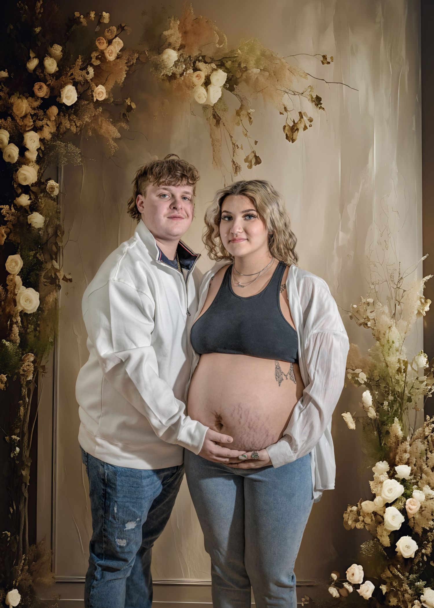 A couple poses together in front of white roses and dried flowers for a maternity photo shoot.