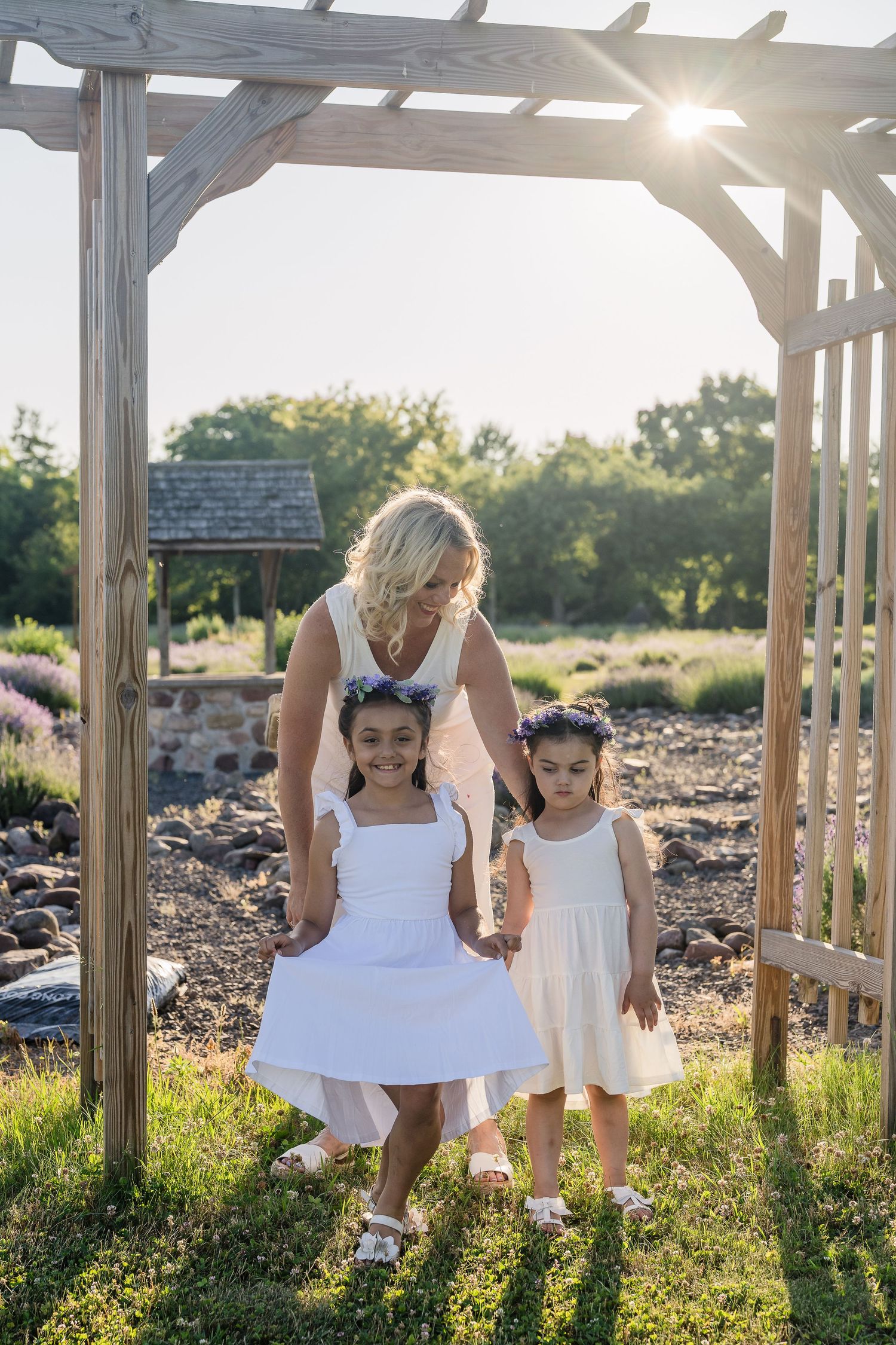 Children in white dresses dance and play under a wooden pergola during a sunny summer evening. Family photography.