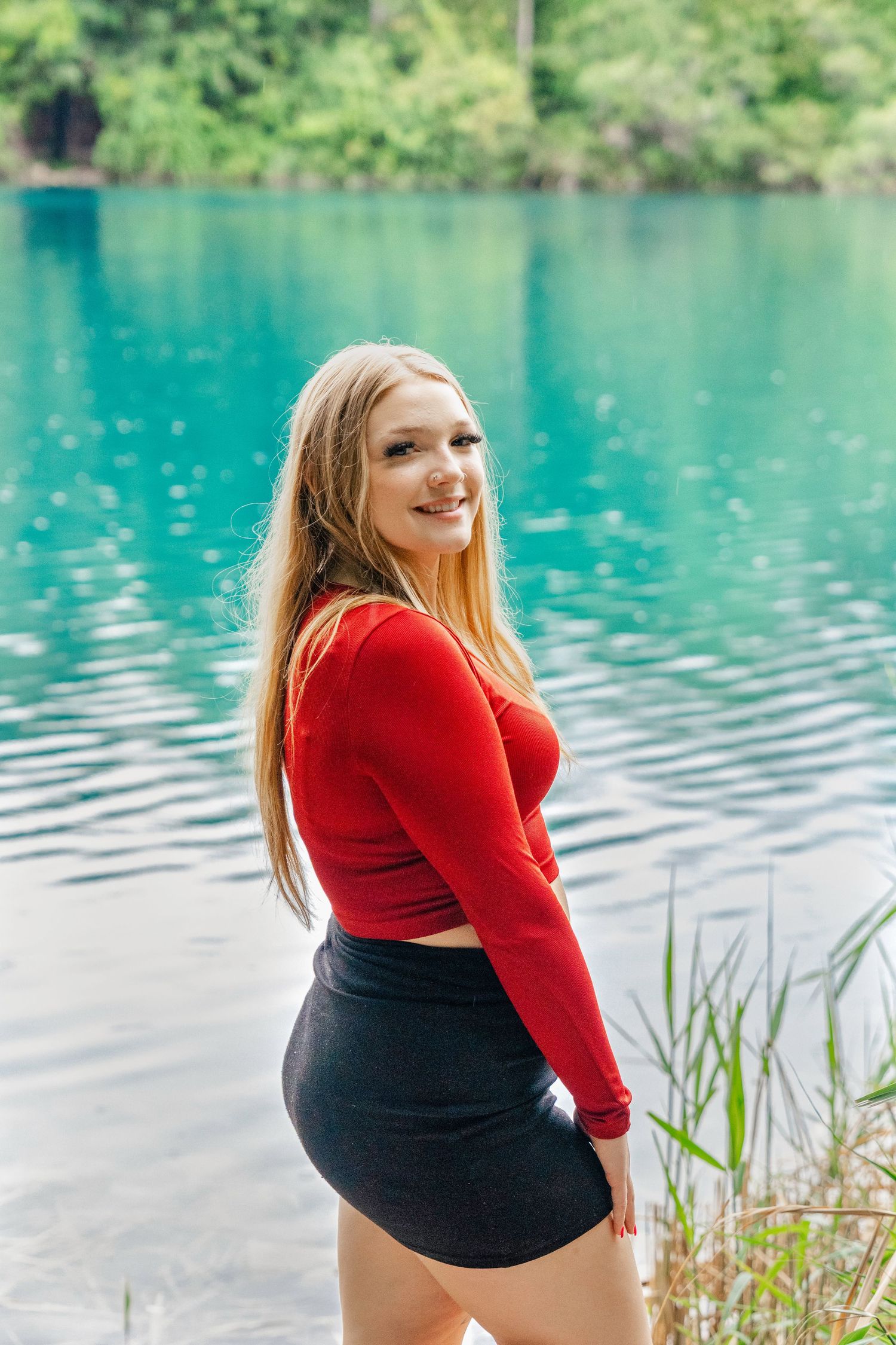 A woman in a red sweater and black shorts poses by a serene turquoise lake surrounded by greenery. Green lakes state park