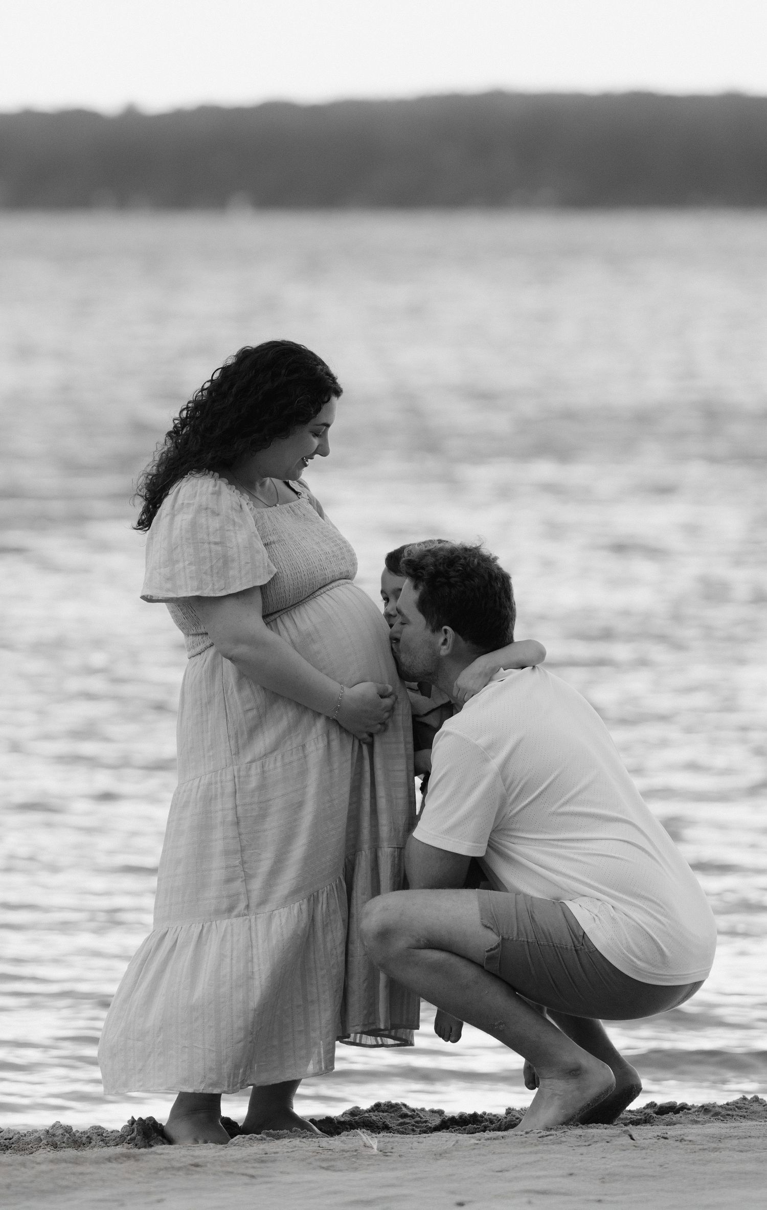 A tender black and white beach scene of an expectant couple sharing an intimate moment by the water.