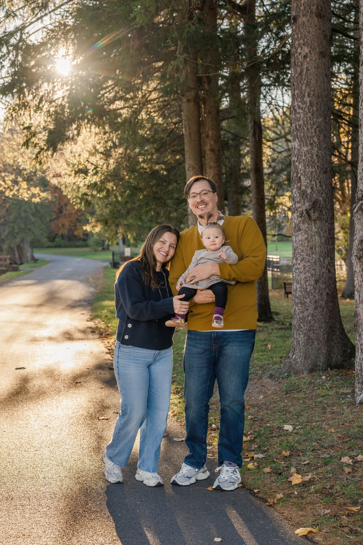 Family walking together on a sunlit autumn path surrounded by tall pine trees.