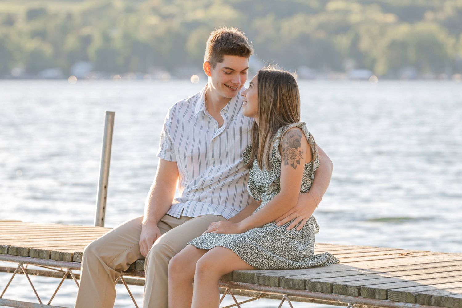 A couple shares a romantic moment sitting together on a wooden dock by a serene lake at sunset.