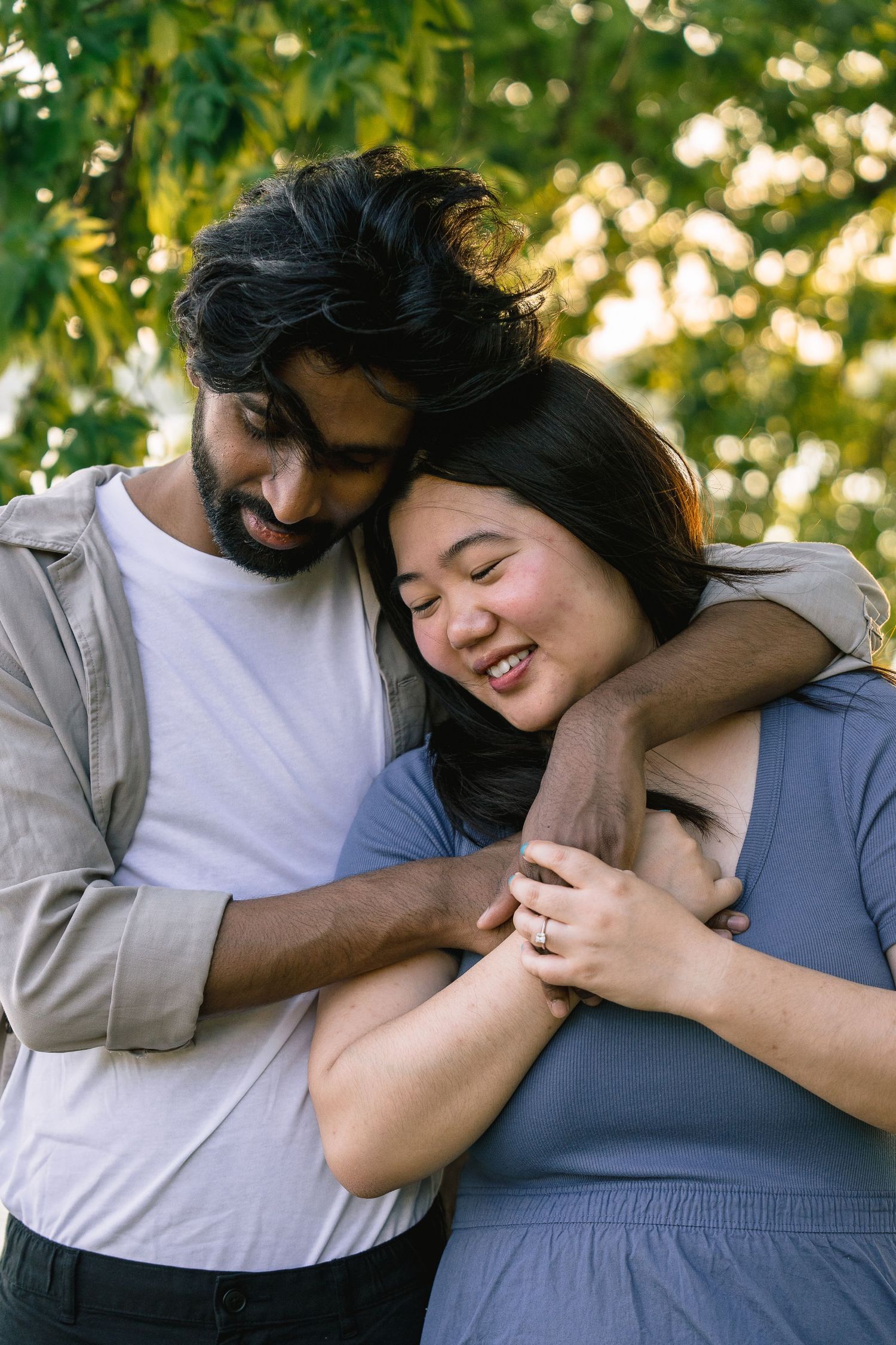 A couple embraces tenderly outdoors with soft bokeh lights and greenery in the background.
