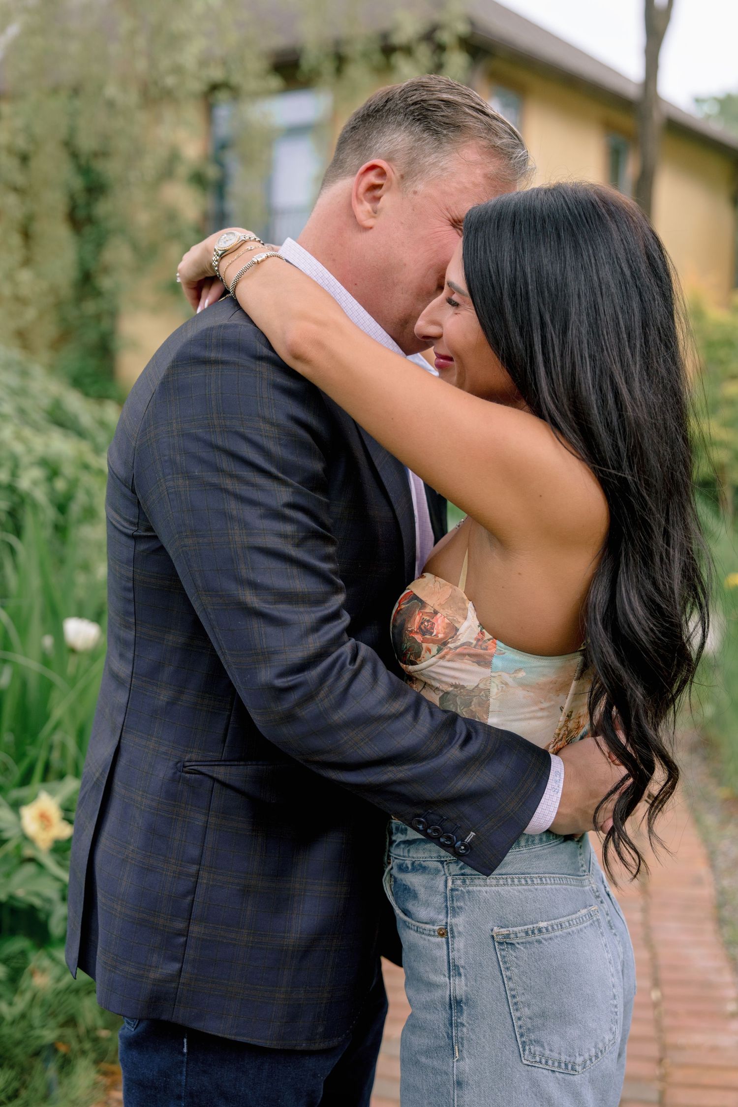A romantic couple embraces in front of a yellow house surrounded by garden foliage.