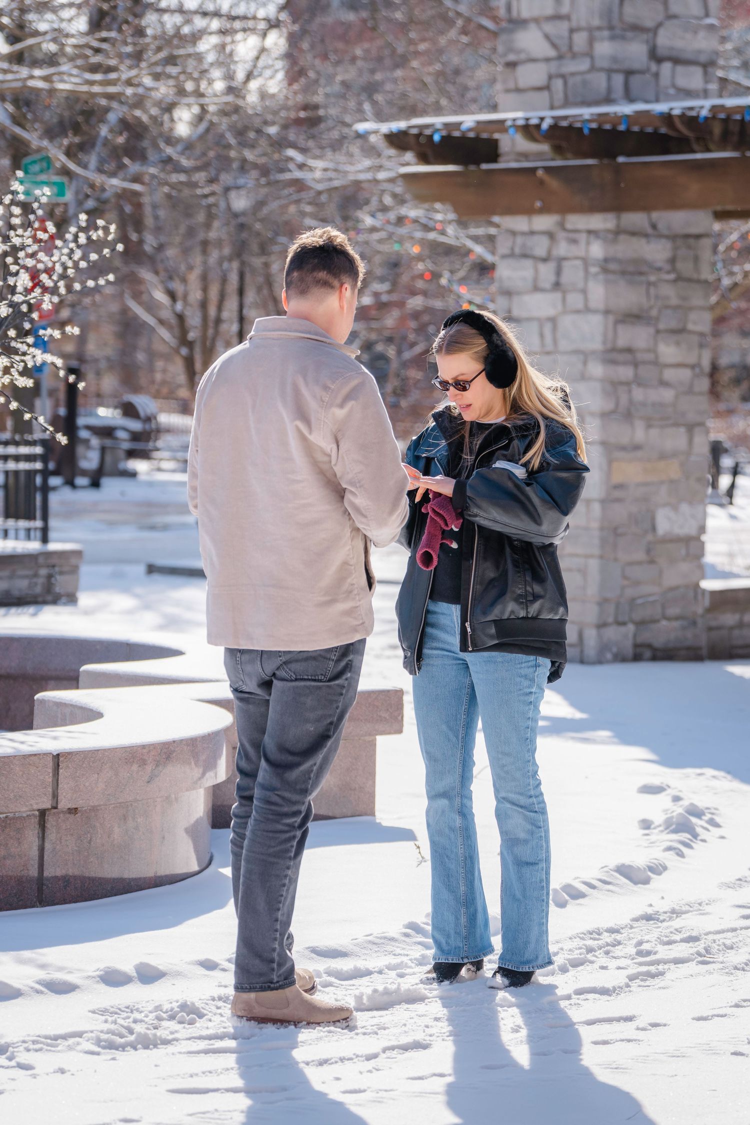 Couple standing in snowy outdoor plaza, woman in black jacket and ear muffs looking at phone, winter day.