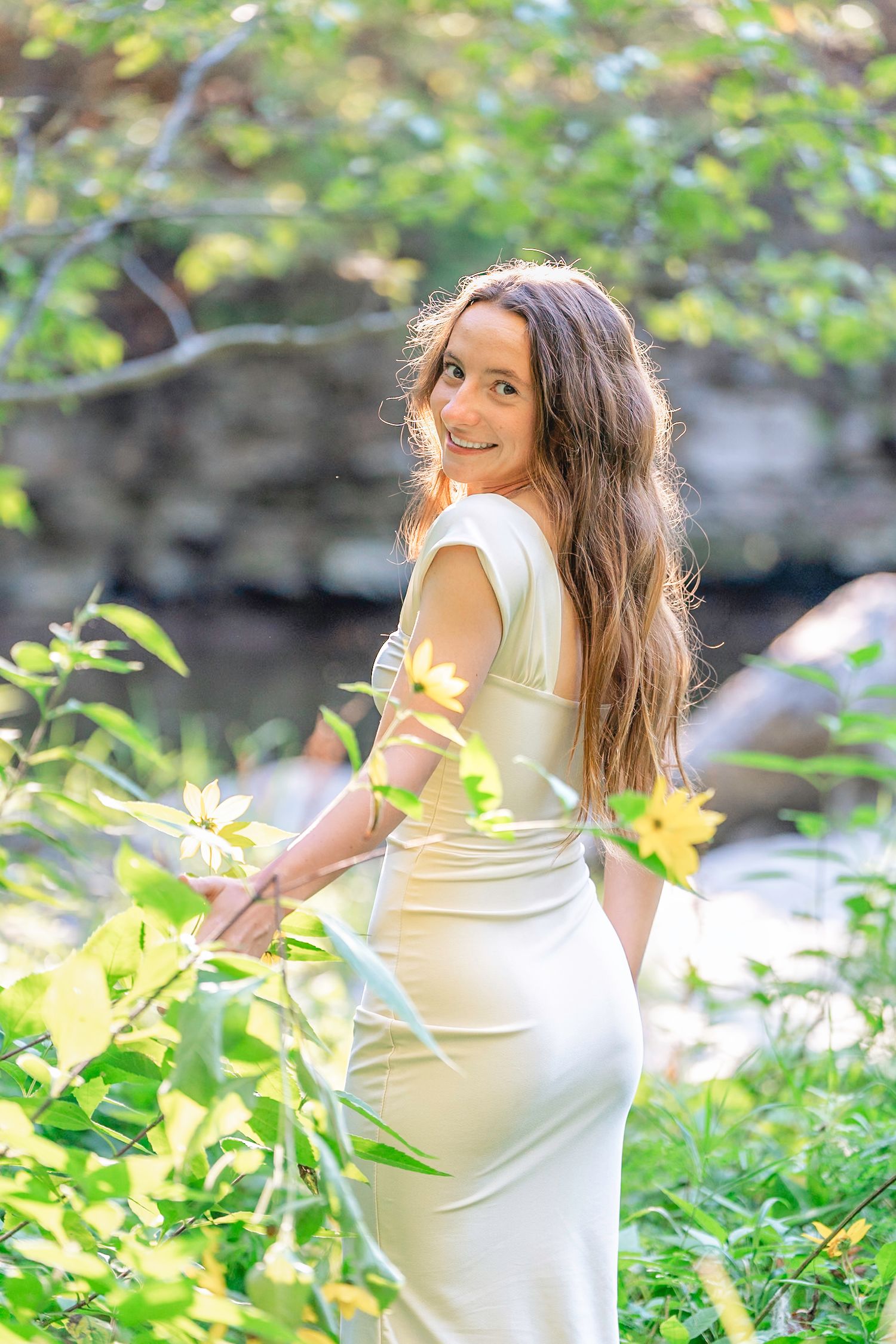 Woman in white dress poses gracefully in a sunlit garden setting with green foliage in the background.