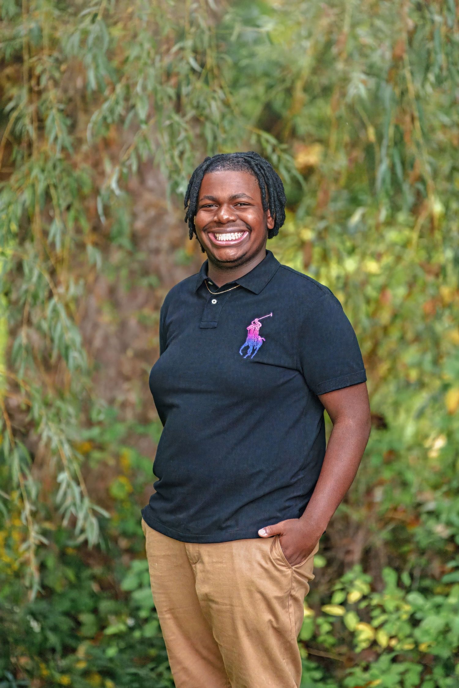 A smiling person wearing a black polo shirt with a pink logo stands outdoors against a backdrop of willow trees.