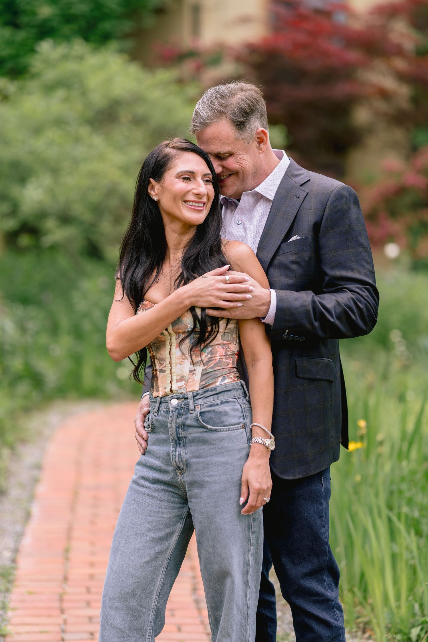 A smiling couple embraces on a brick garden path, woman in floral top and jeans, man in dark suit, surrounded by lush greenery.