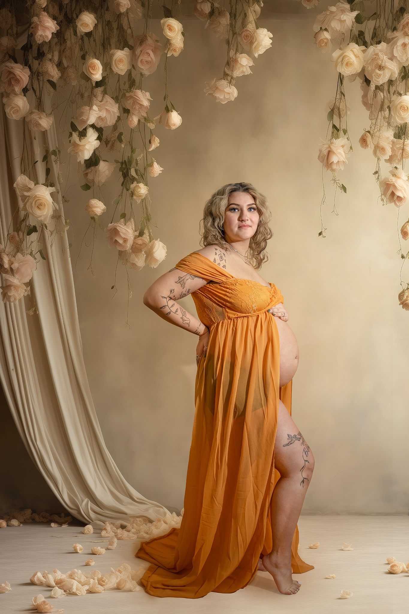 A maternity photo featuring a flowing orange dress against a romantic backdrop with hanging cream-colored roses and draping fabric.