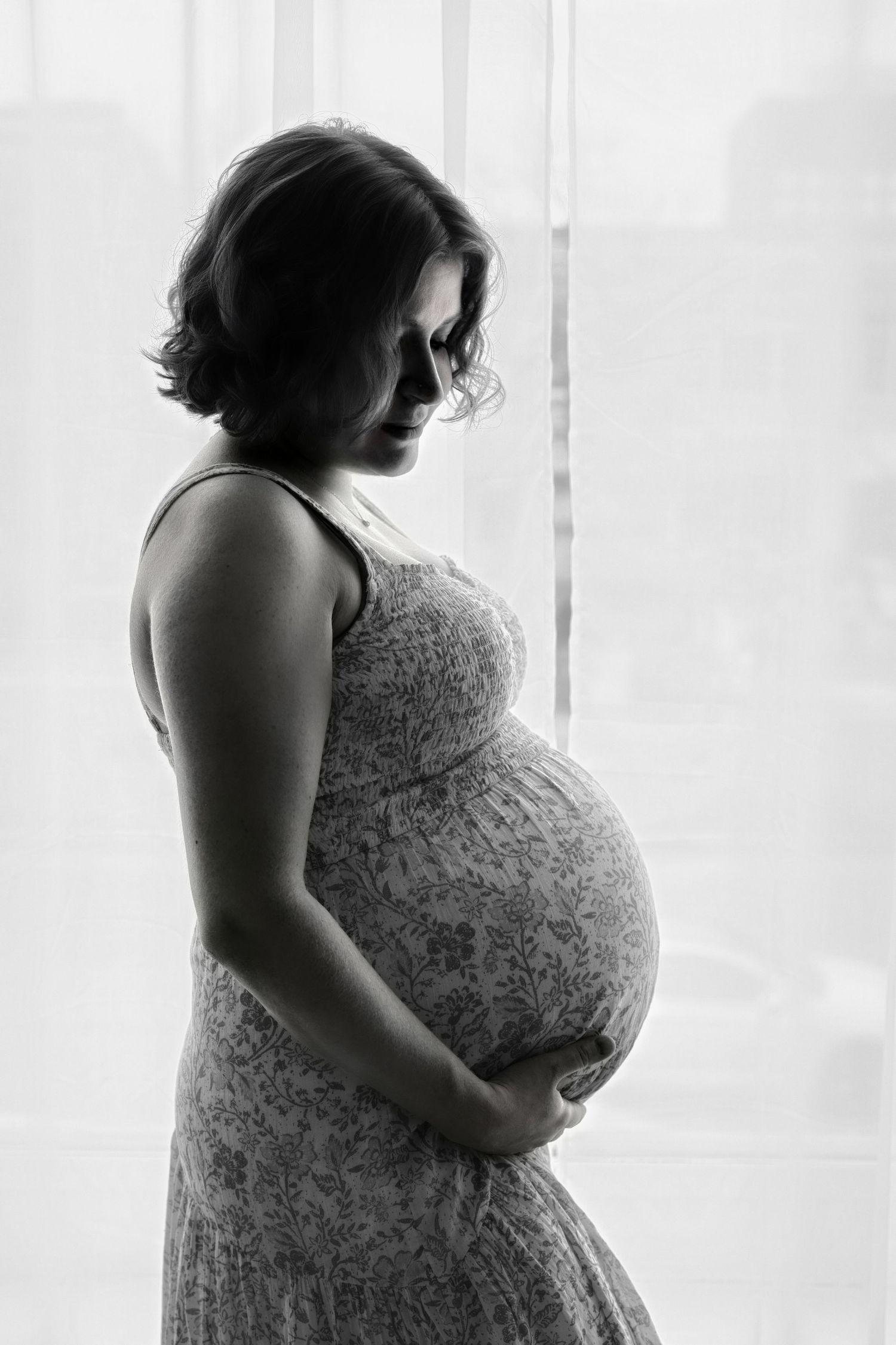 Artistic black and white maternity photo of an expectant mother in a lace dress by a window.
